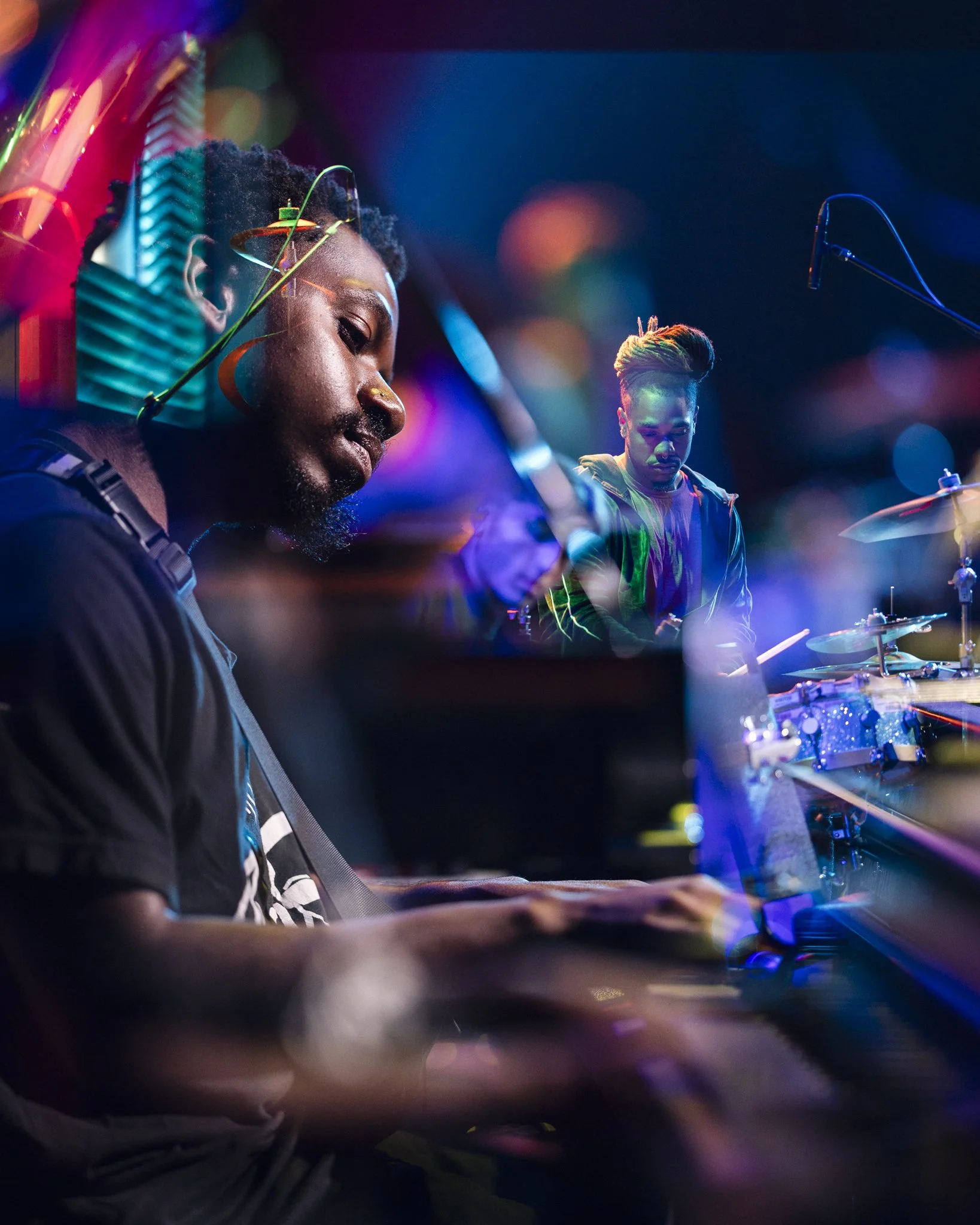 Two musicians performing on stage with colorful lights, one playing the piano with headphones and the other playing drums, atmospheric and vibrant concert scene.