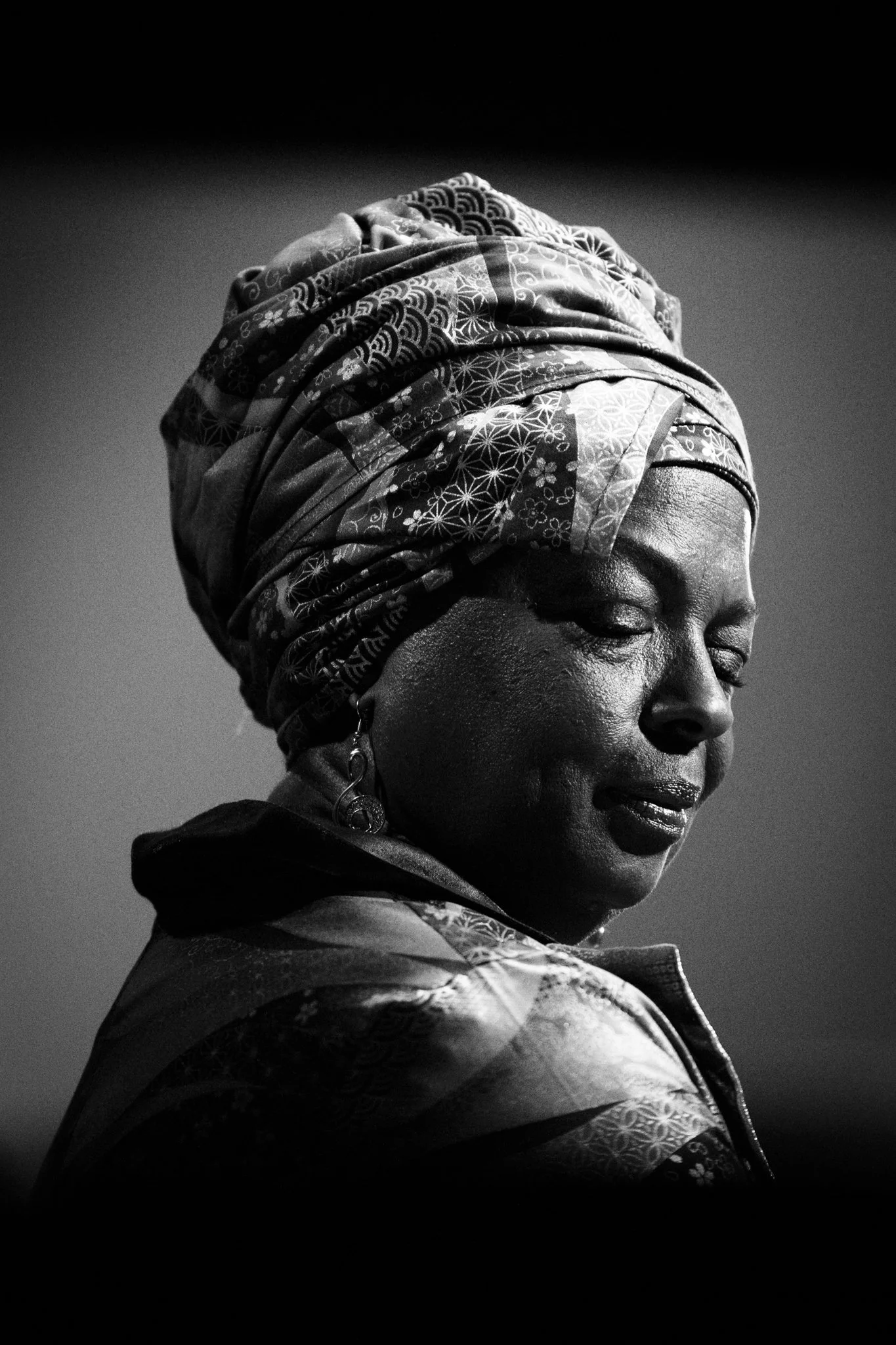 A black and white portrait of a woman wearing a patterned headscarf and earrings, with her eyes closed and a serene expression.