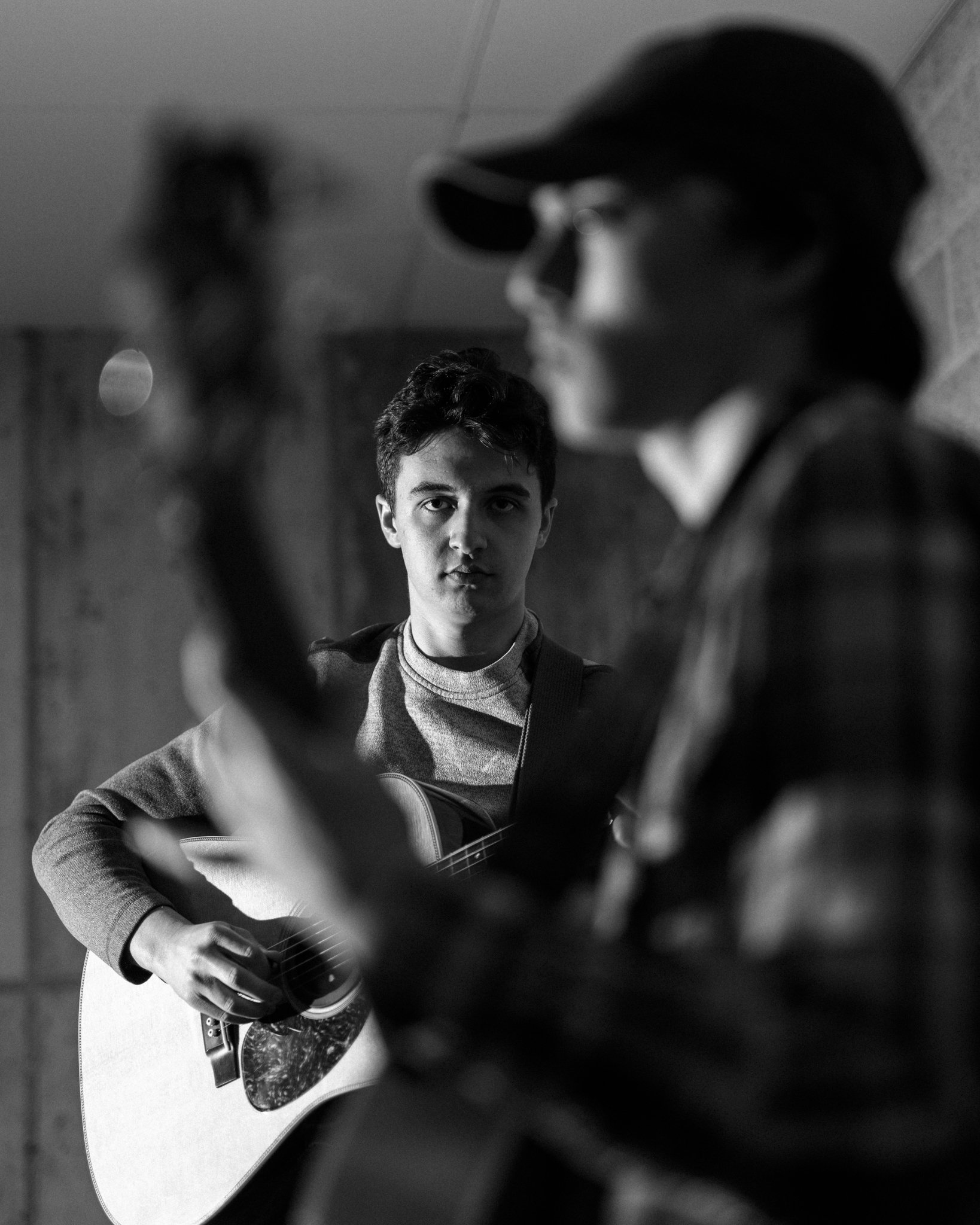 A young man is playing an acoustic guitar while another person in the foreground, wearing a cap and glasses, is also playing a guitar. The scene is in black and white.