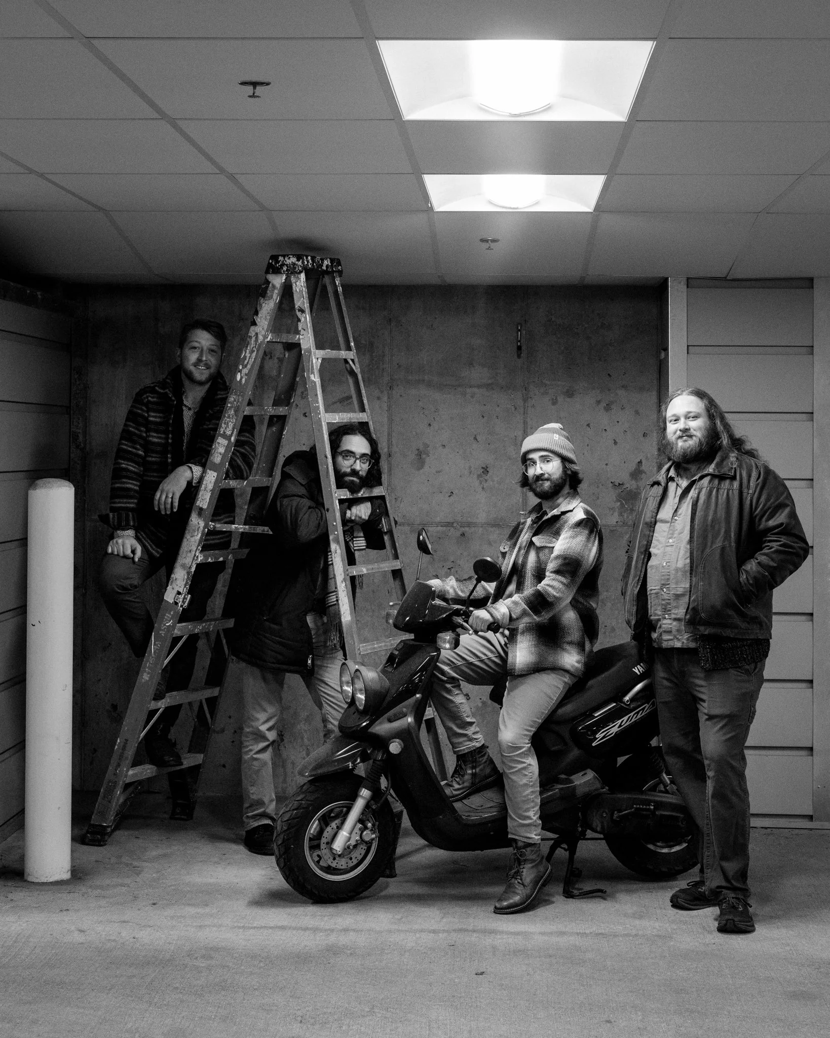 Black and white photo of four men in a garage. One is sitting on a scooter, while another is standing next to it. One man is sitting on a ladder, and another stands to the right, all looking at the camera.