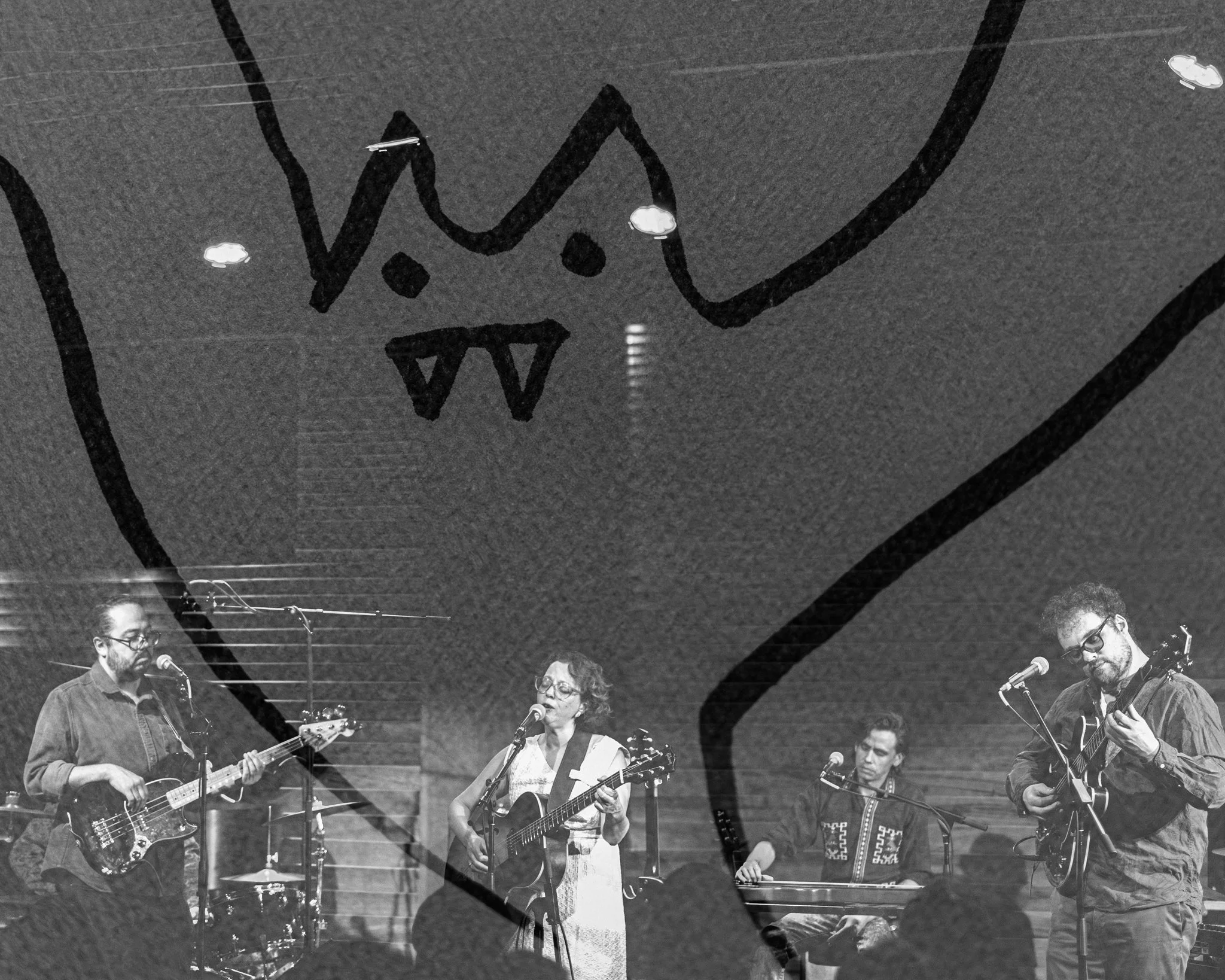 A black and white image of a band performing on stage, with four musicians playing guitars, drums, and keyboard, under a large drawing of a monster face with sharp teeth and pointed features projected onto the backdrop.