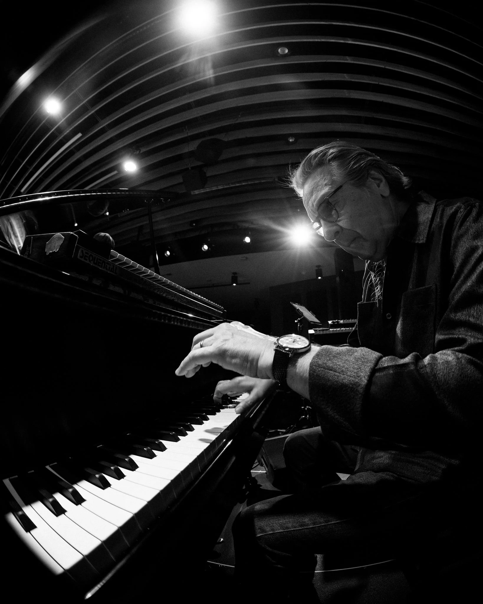 A black and white photo of an older man playing a grand piano in a music rehearsal space, with ambient lighting and an arched wooden ceiling.