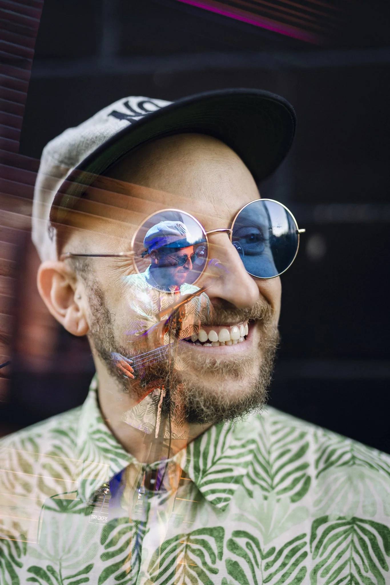 A smiling man wearing round sunglasses, a baseball cap, and a patterned shirt, with his reflection visible in the sunglasses.