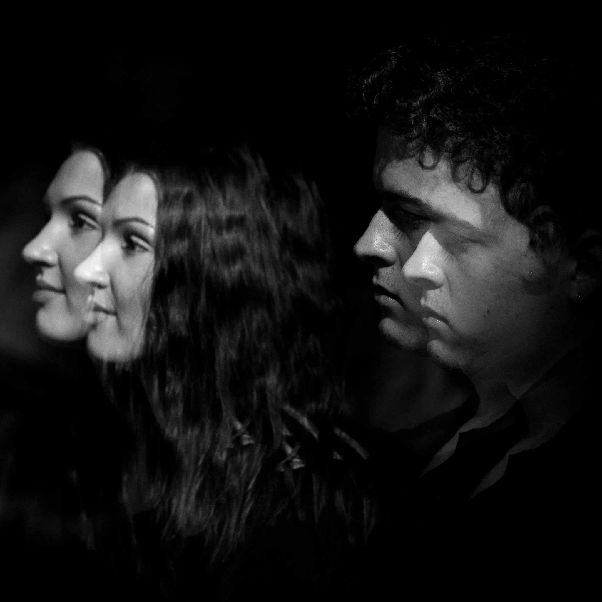 Side view of three young adults, two women and one man, facing to the left, with a dark background, in black and white photo.