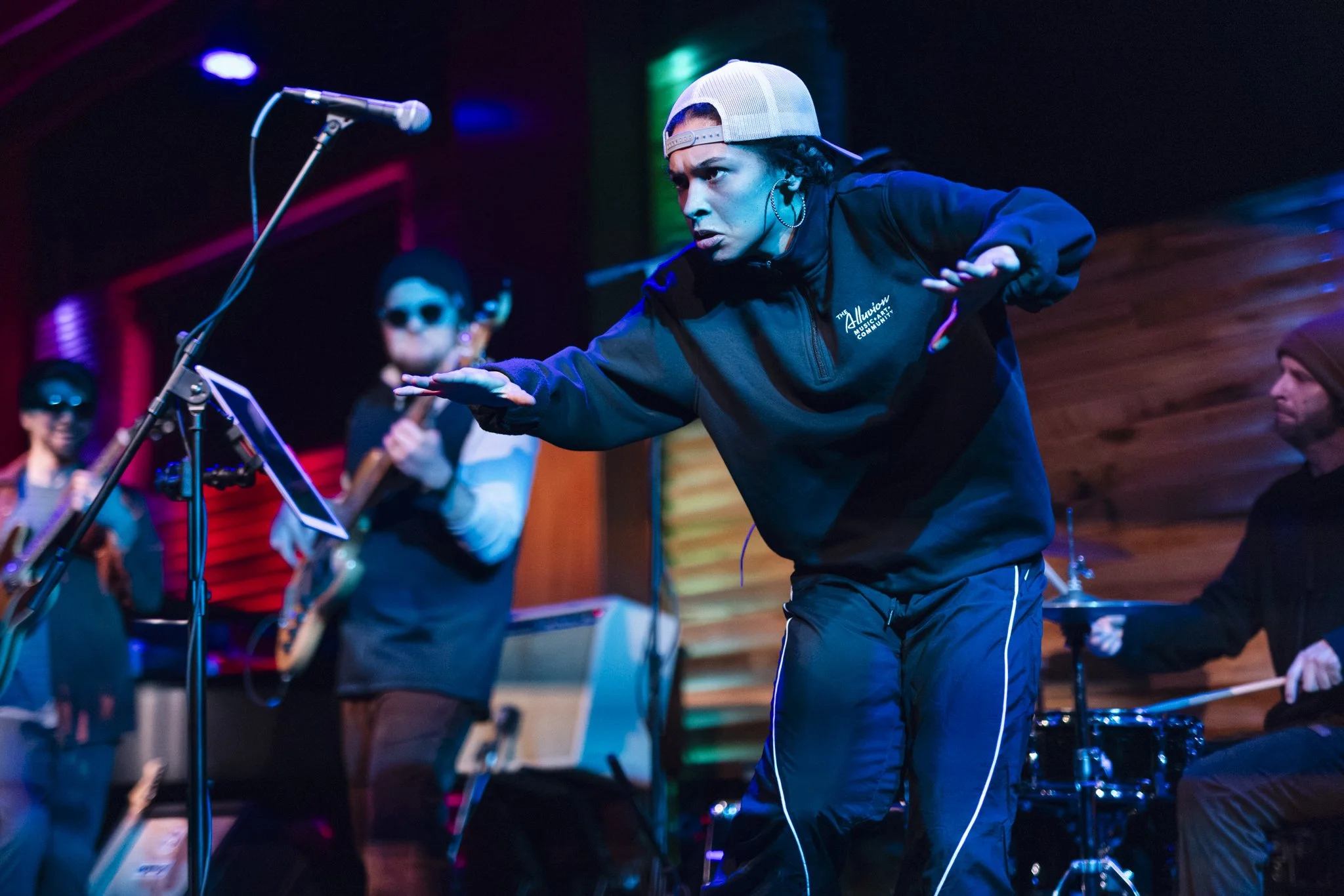 A person in a baseball cap and black hoodie is dancing on stage with a band, with two guitar players and a drummer in the background, colorful stage lighting.