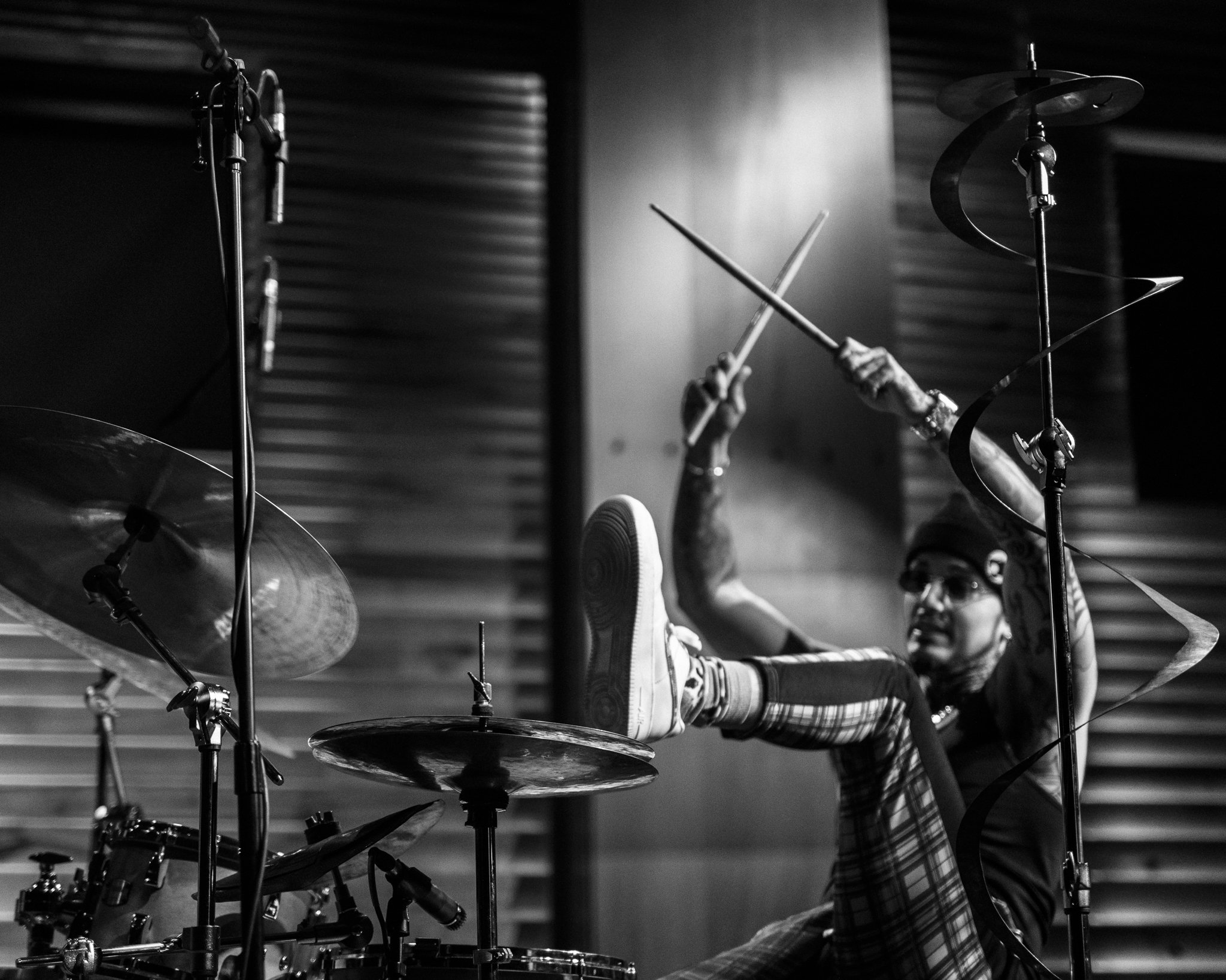 A person wearing glasses and a hat, sitting behind a drum kit, with one foot raised on a cymbal, holding drumsticks in the air, in a recording studio.