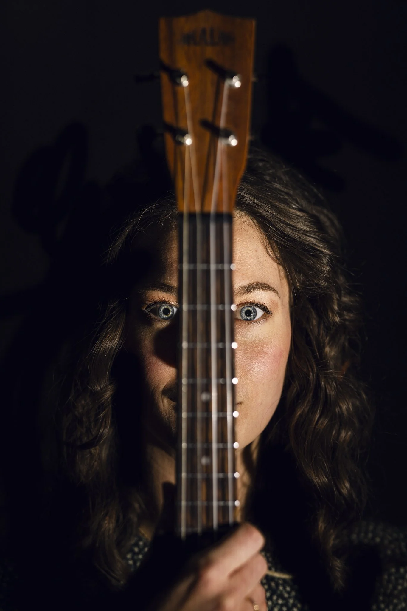 A woman with curly brown hair and blue eyes holding a ukulele vertically in front of her face, partially obscuring it, against a dark background.