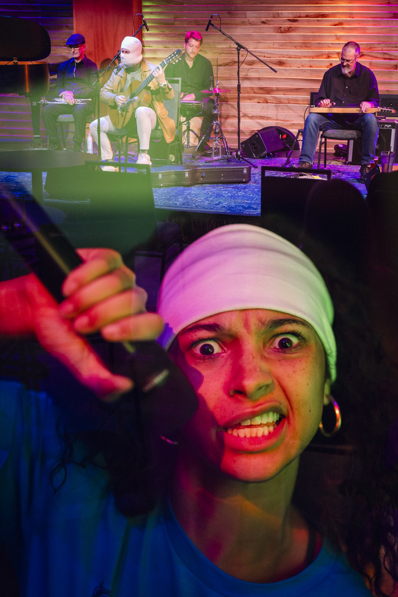 A woman with a white headscarf and hoop earrings looks angry and is holding a stick, with a music performance happening in the background on a stage with wooden panels, featuring four musicians playing various instruments.