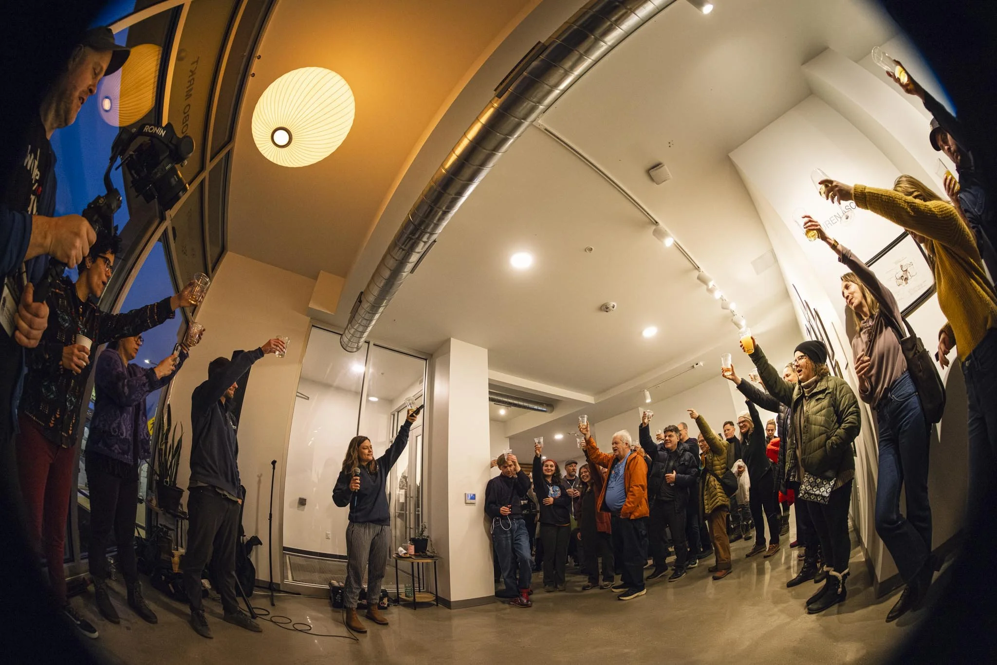 A group of people celebrate at a gathering in a modern indoor space, raising their glasses in a toast. The room has warm lighting, a white ceiling, and is decorated with framed artwork.