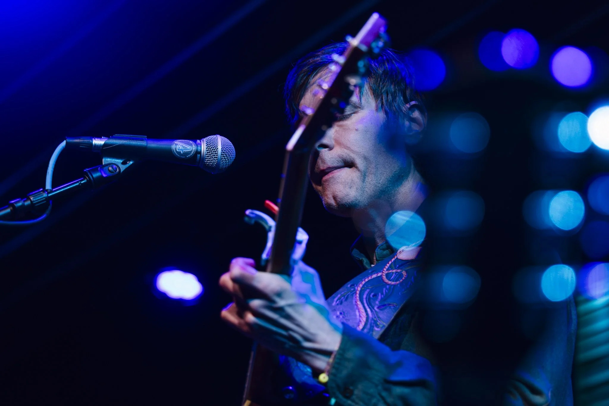 A male musician playing an acoustic guitar on stage, singing into a microphone with blue and purple stage lighting and bokeh effects.