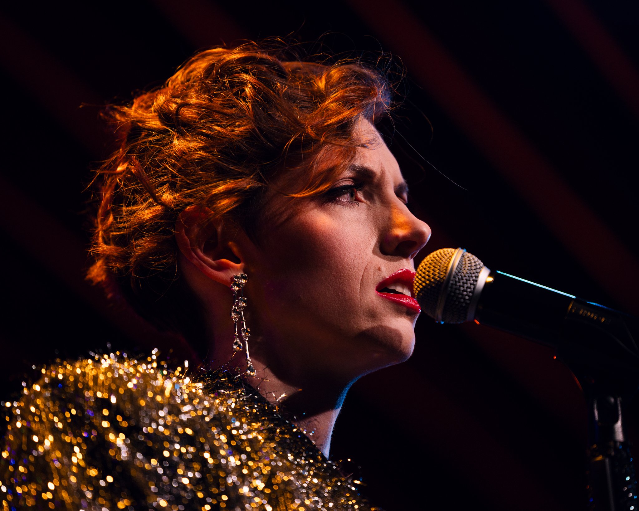 A woman with short, curly hair singing into a microphone, wearing eye-catching earrings and a gold sequin outfit.
