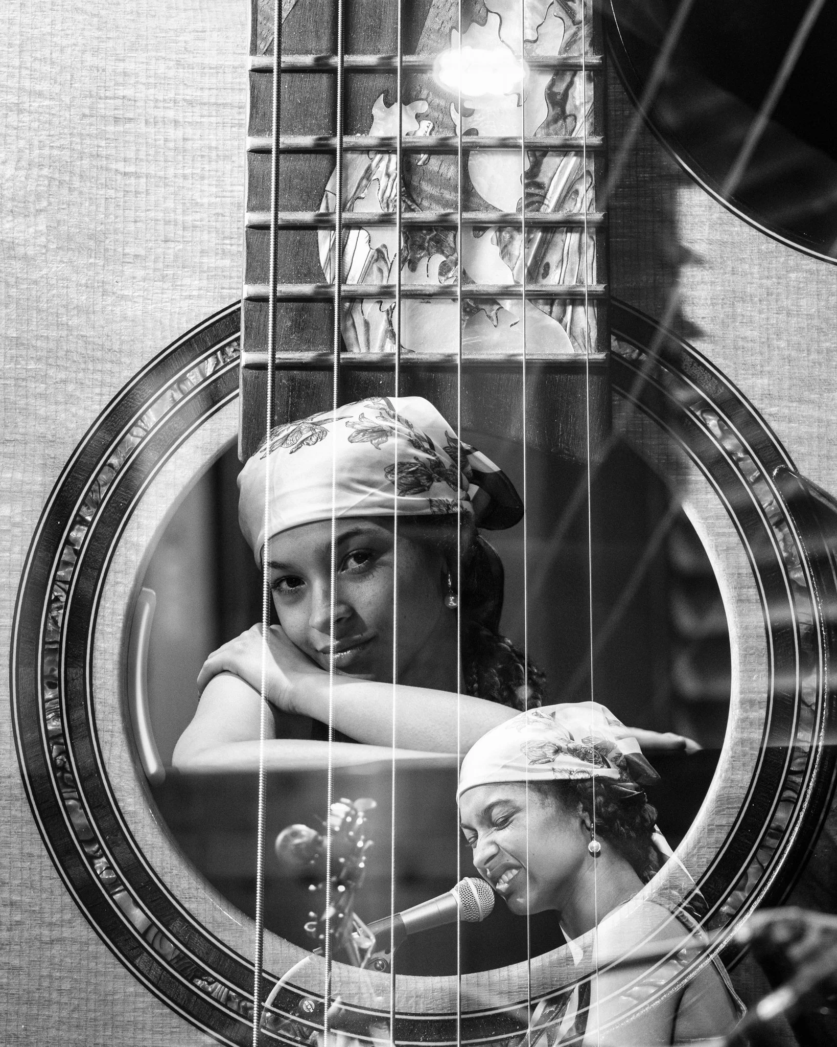 Black and white photo of a woman looking through a guitar's soundhole, with her reflection visible in the guitar's interior. The woman is wearing a headscarf and earrings, and appears to be singing into a microphone.