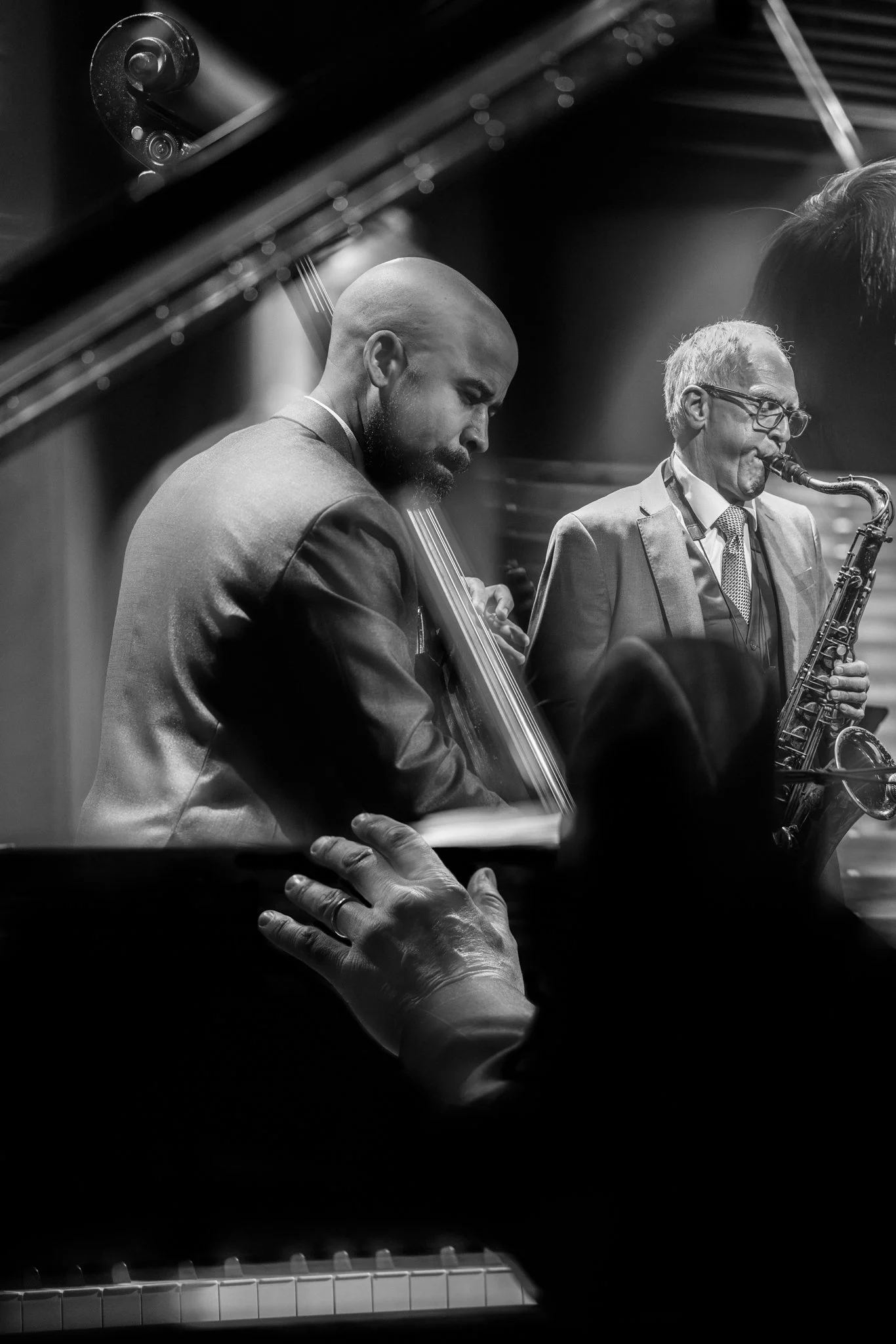 Black and white photo of three jazz musicians performing, including a pianist, a bassist, and a saxophonist, with a hand of an audience member visible in the foreground.