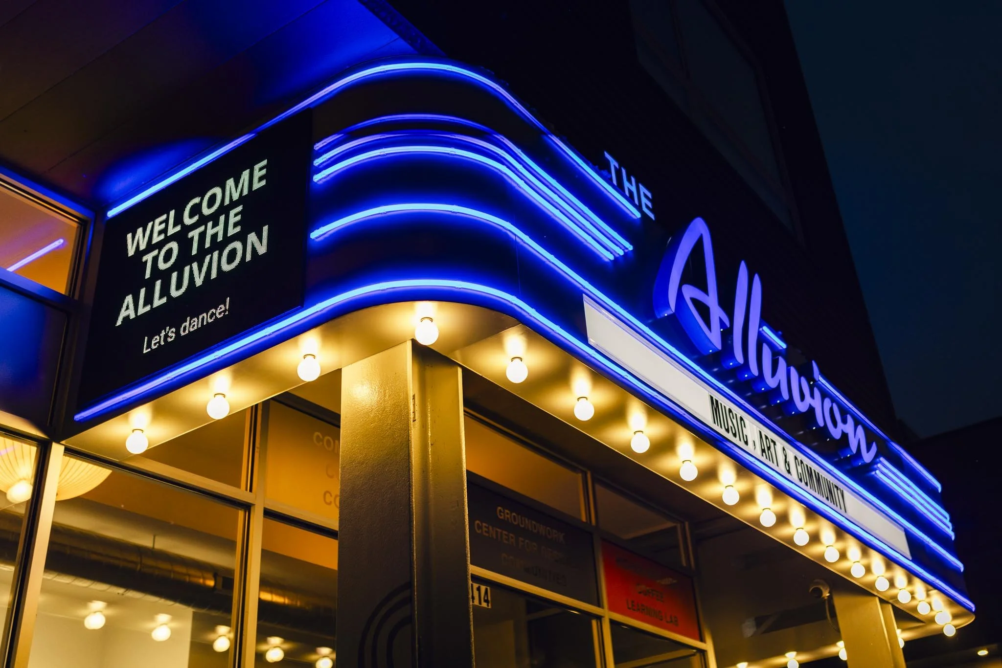 Night view of the Alluvion entertainment venue's illuminated marquee with blue neon lights, a sign welcoming guests, and warm yellow light bulbs outlining the entrance.