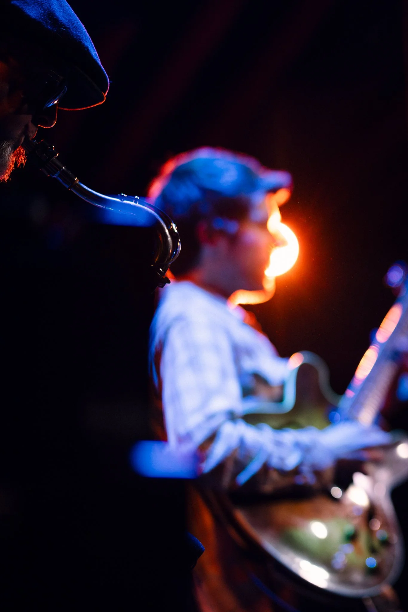 Musician playing the guitar with a bright, warm light in the background, and a saxophone player partially visible in the foreground.