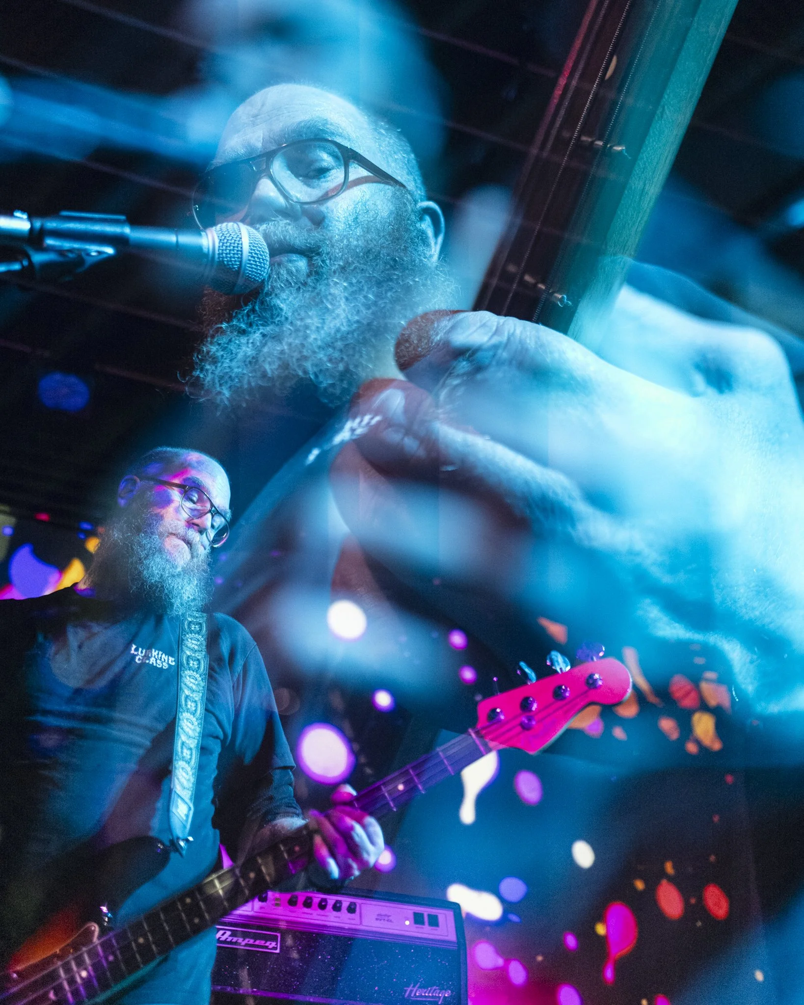 Bearded man with glasses playing electric guitar and singing into a microphone on stage with colorful lights.