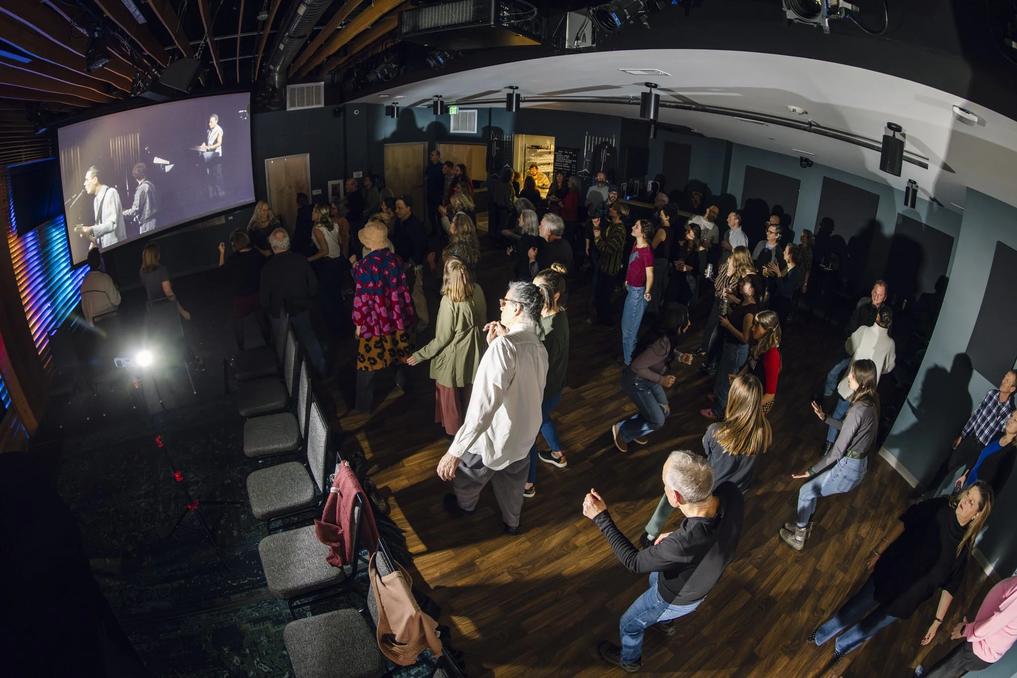 People dancing and socializing in a dark indoor event space with a large screen displaying a group of performers.