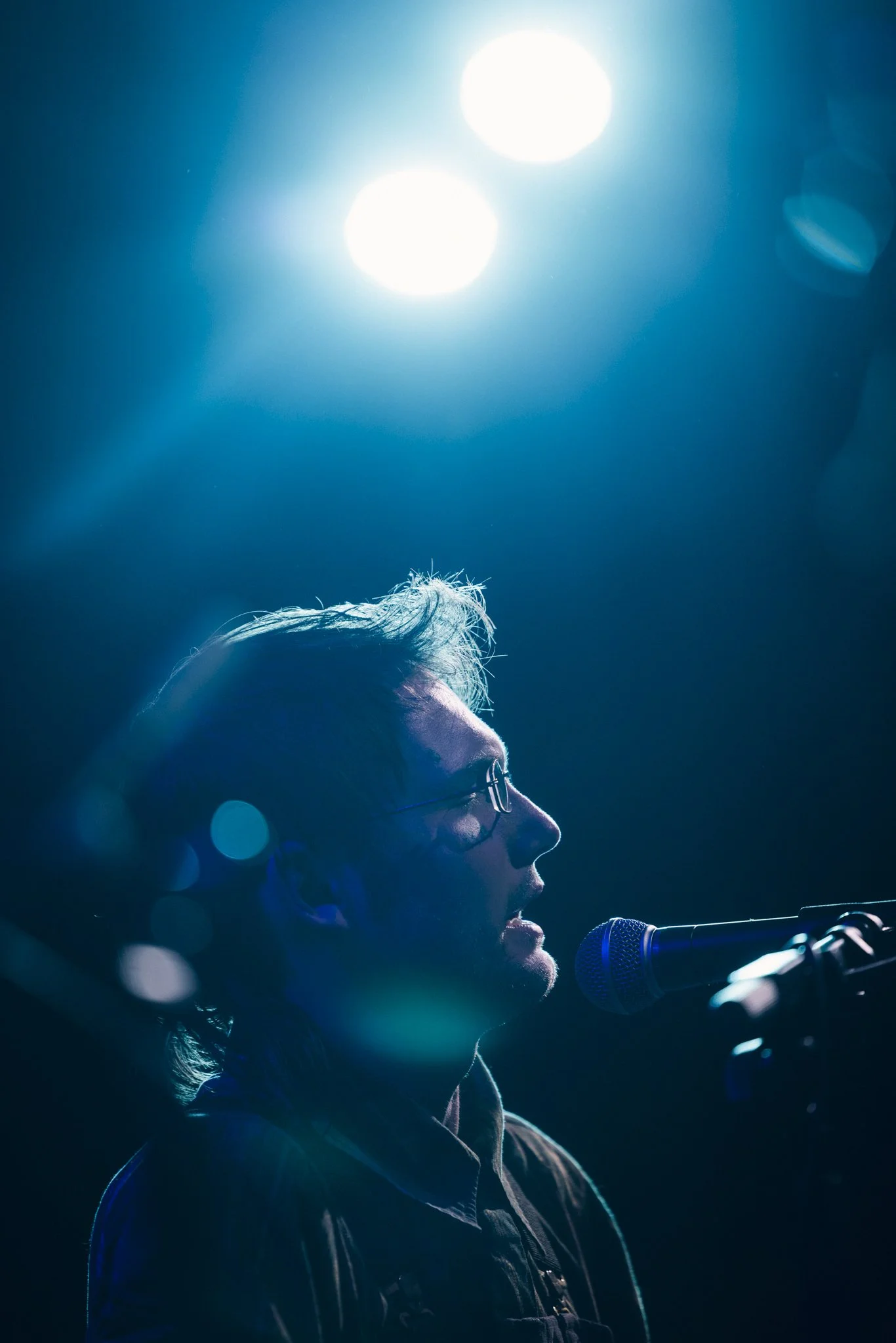 A man with glasses singing or speaking into a microphone on stage, illuminated by blue stage lighting.