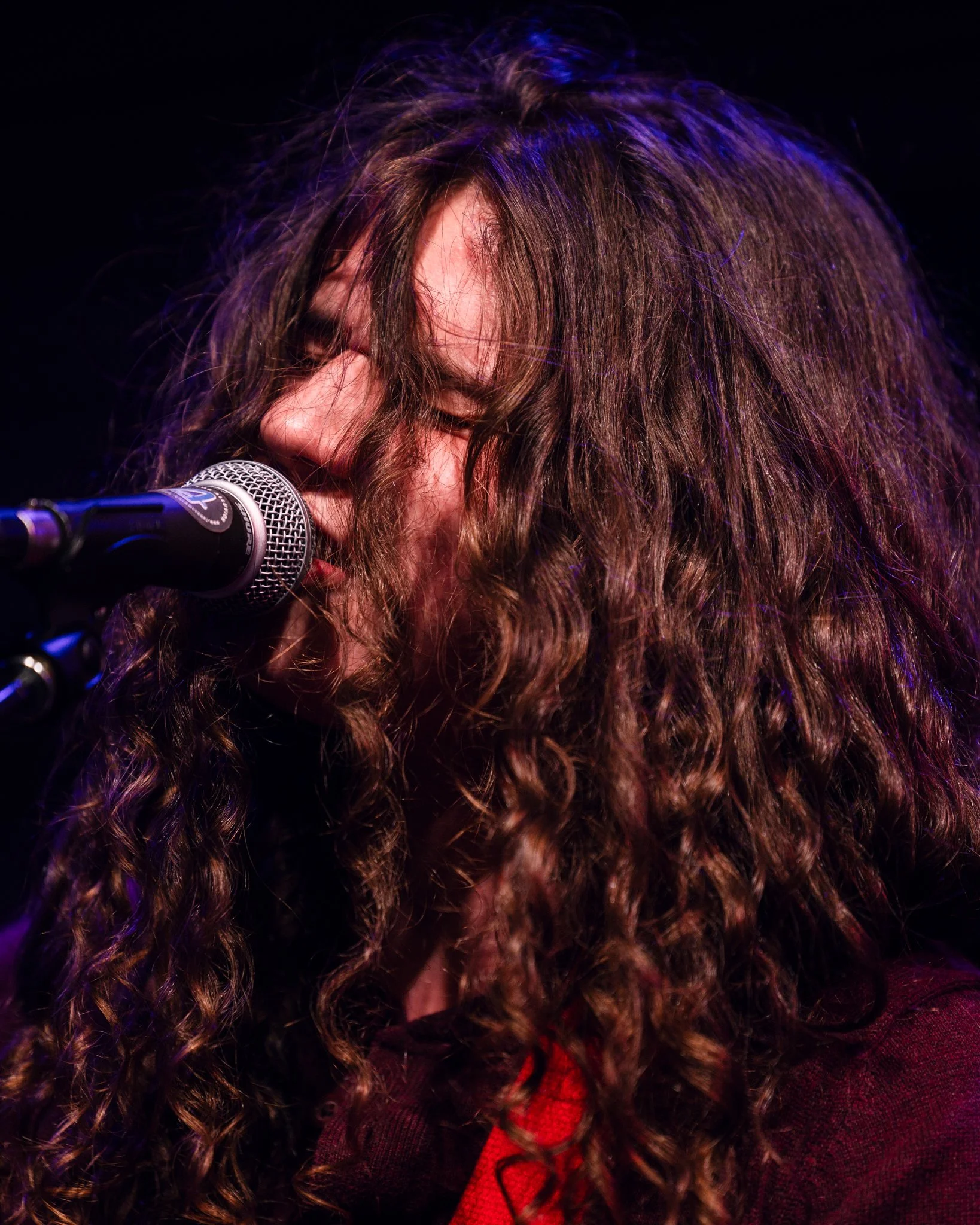 Person with curly hair singing into a microphone, with closed eyes, on a dark stage.