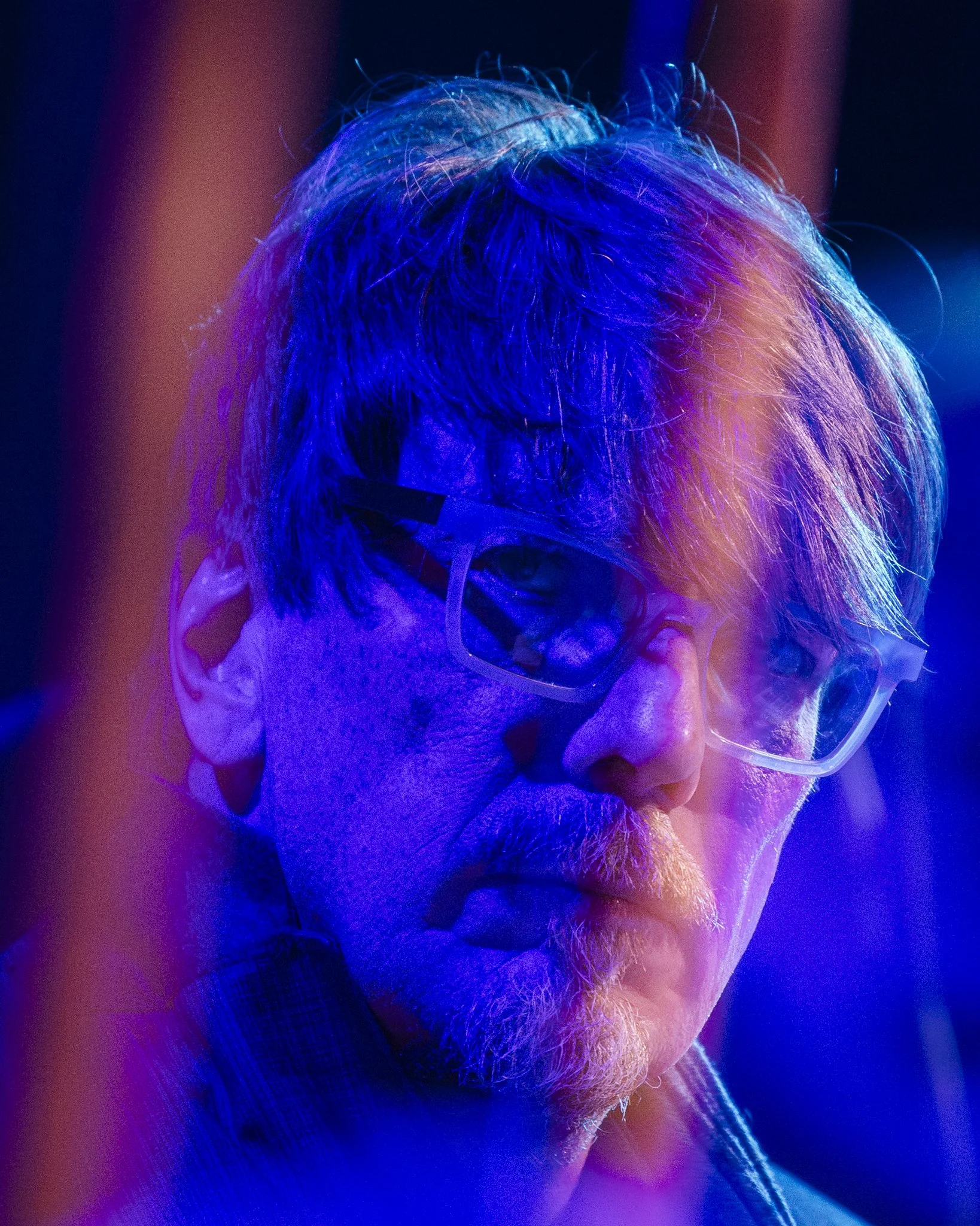 A close-up portrait of a man with glasses and a beard, illuminated by blue and purple lighting.