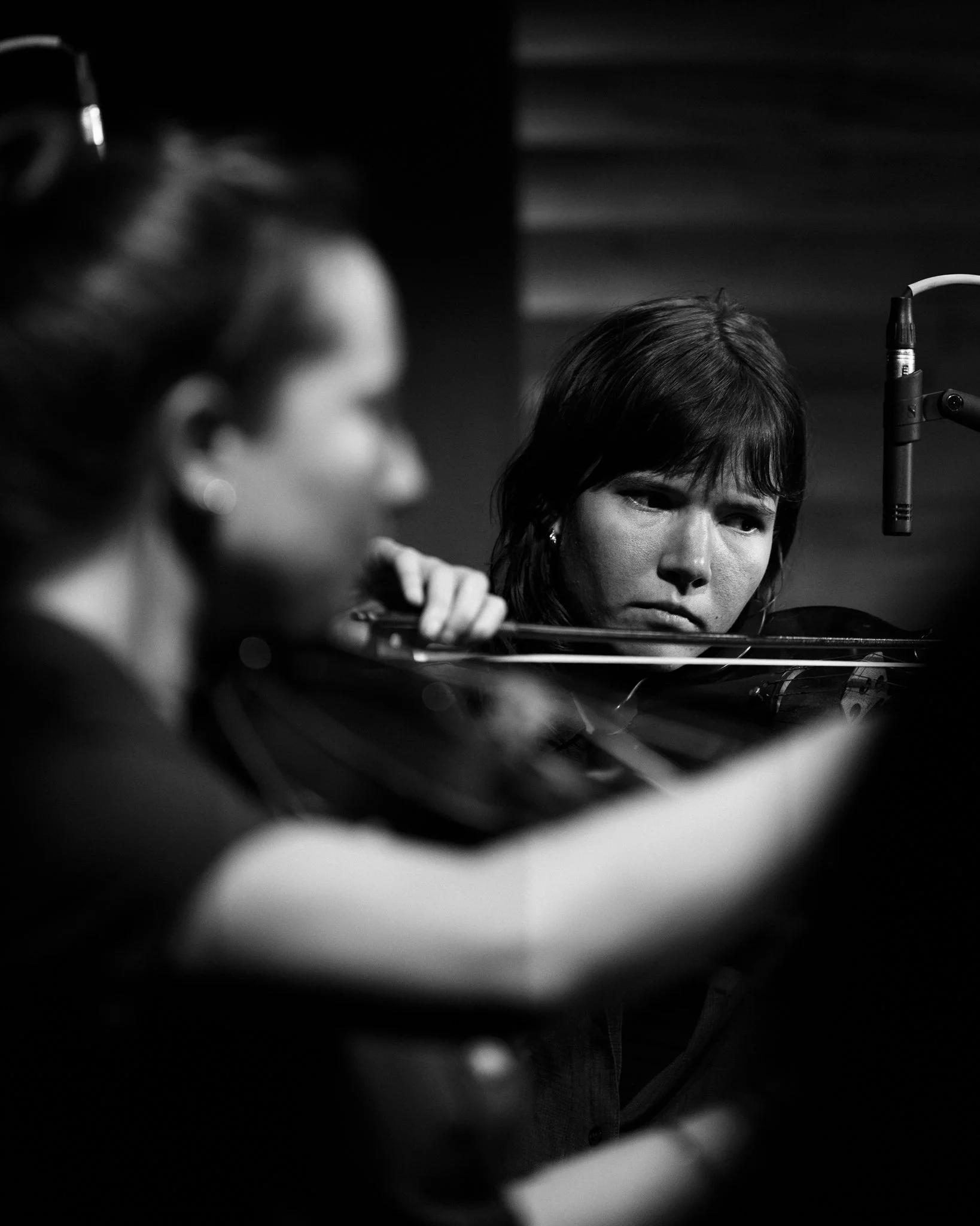 Black and white photo of two women playing violins in a recording studio, one in focus and the other blurred in the foreground.