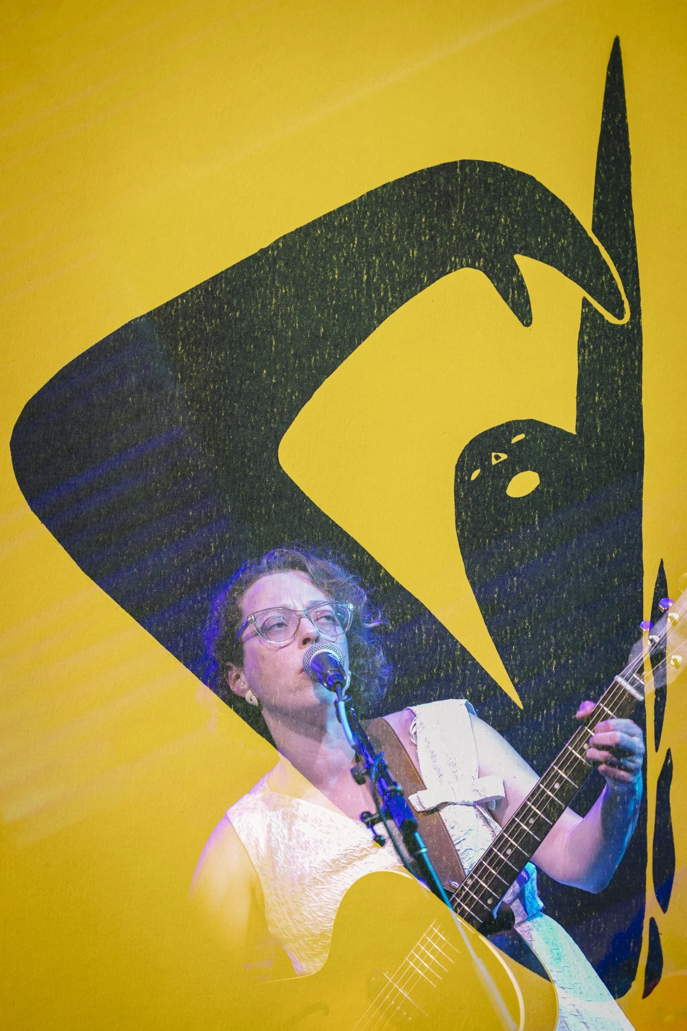 A woman with glasses and curly hair is singing into a microphone while playing an acoustic guitar against a yellow background with a black illustration.