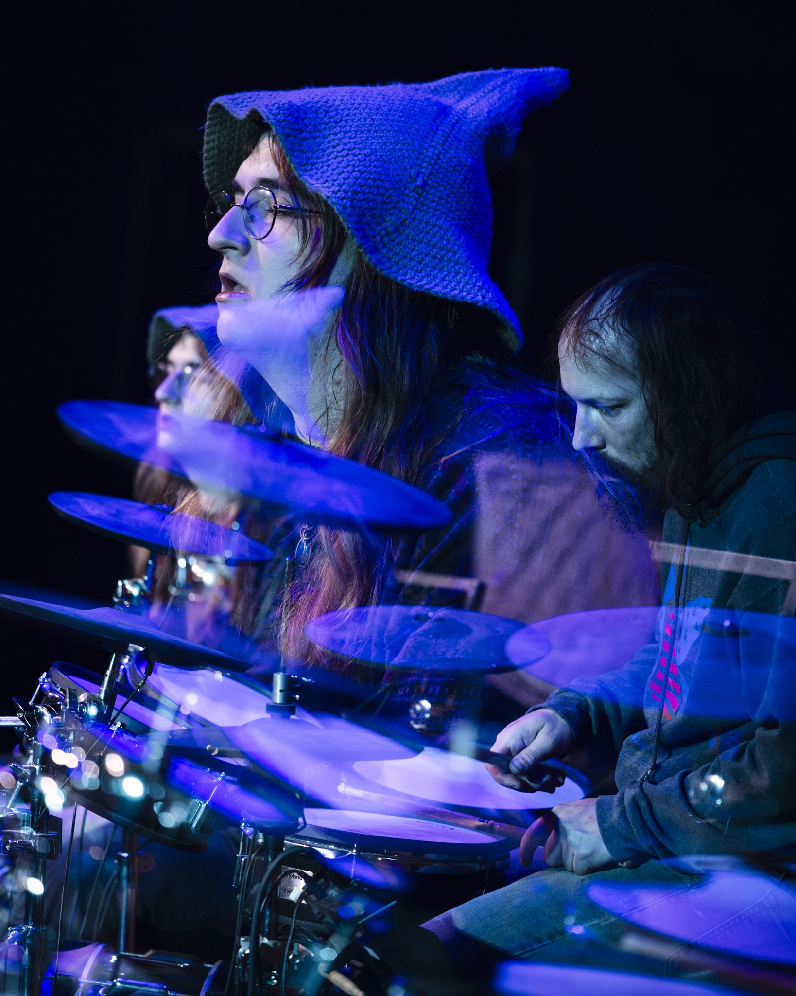 Three musicians performing on stage with blue lighting, one woman wearing glasses and a pointed knit hat playing the drums, two men also playing drums, blurred motion indicates movement.
