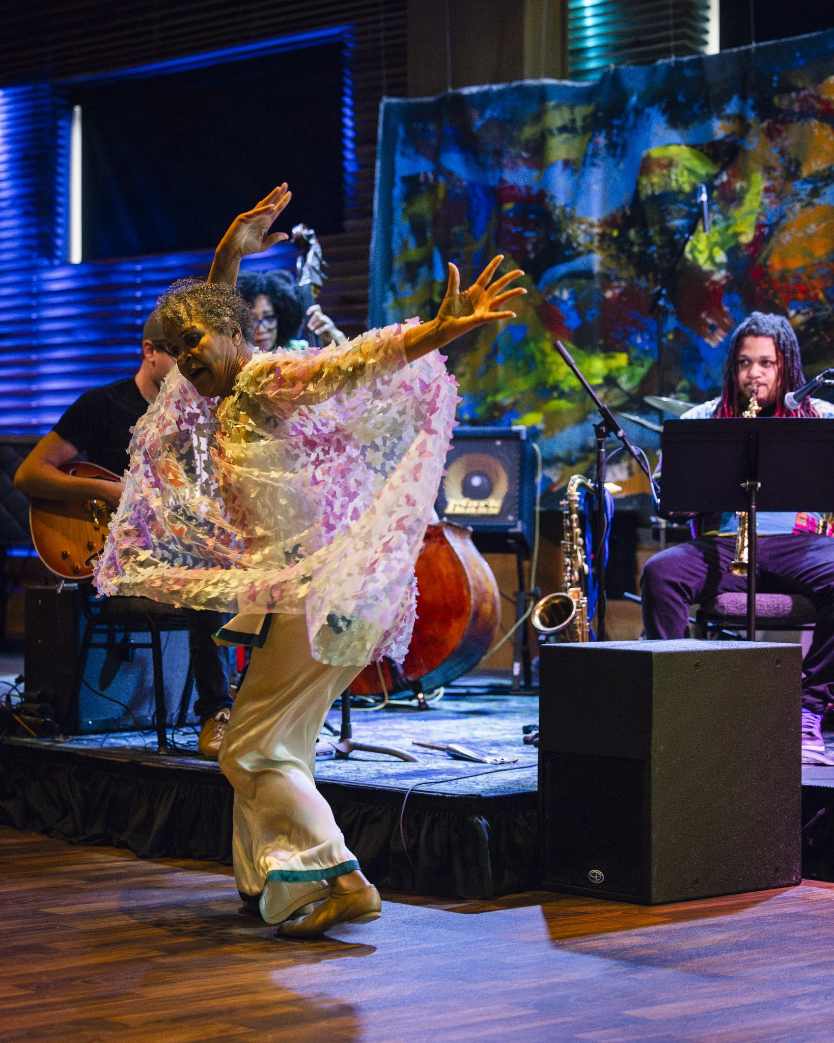 A woman dancing in front of a band performing live music on stage, with a colorful abstract backdrop and various musical instruments.