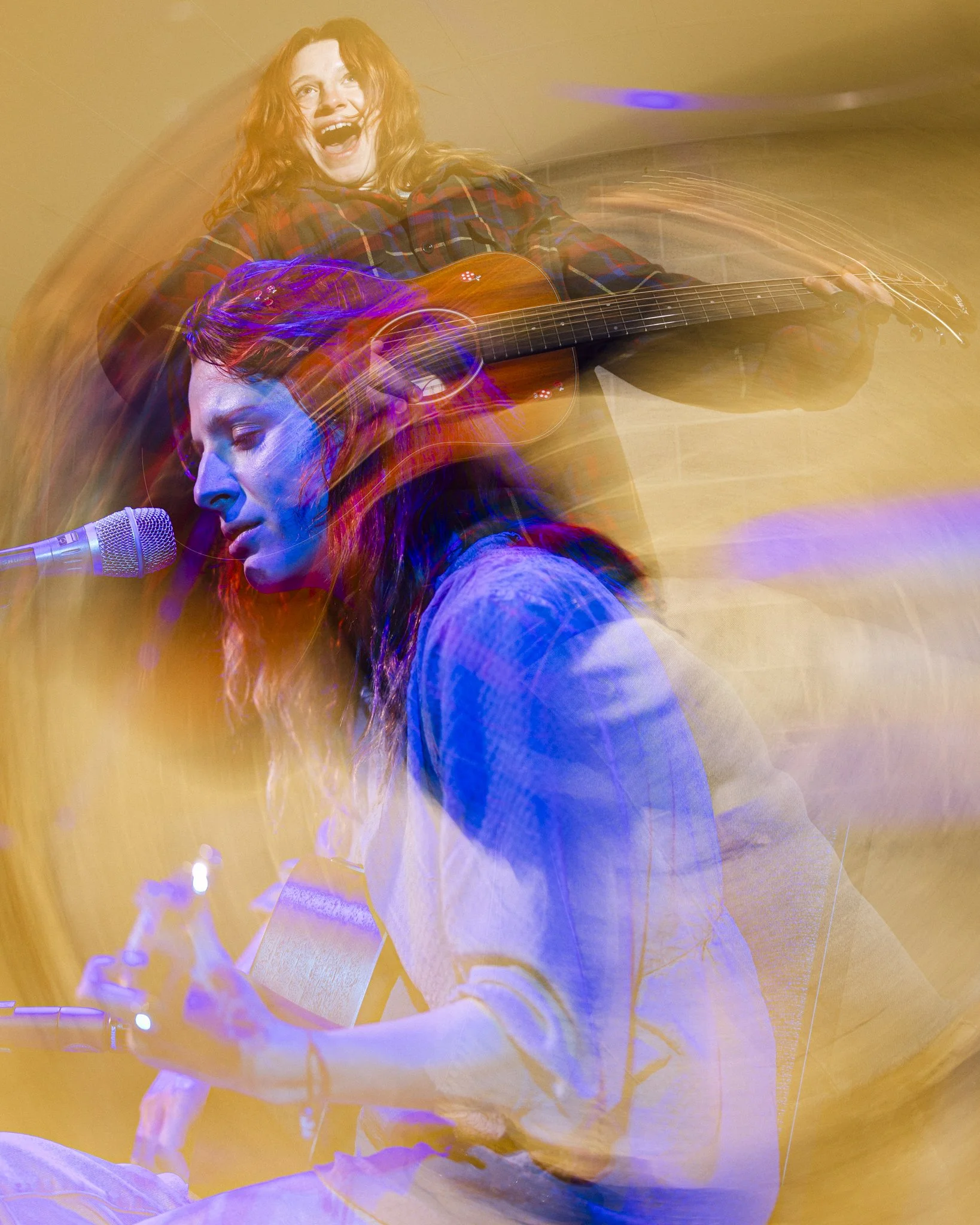 Two women performing music; the woman in the foreground is playing a guitar and singing into a microphone, while the woman in the background is playing an acoustic guitar and singing, with colorful light trails surrounding them.