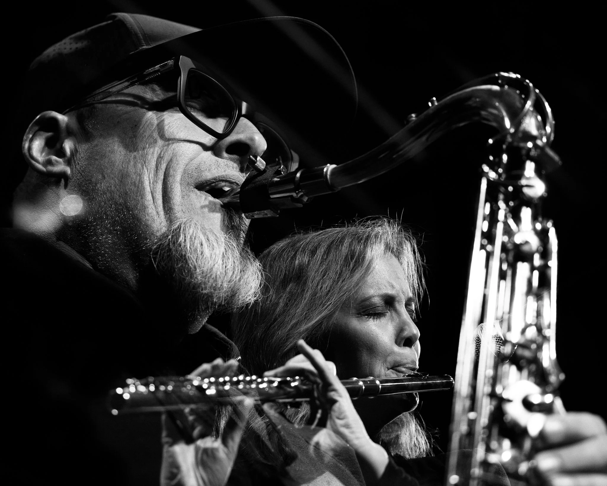 A black and white photo of a man and woman playing flutes, with the man singing into a microphone. The man has glasses and a cap, and the woman has her eyes closed.