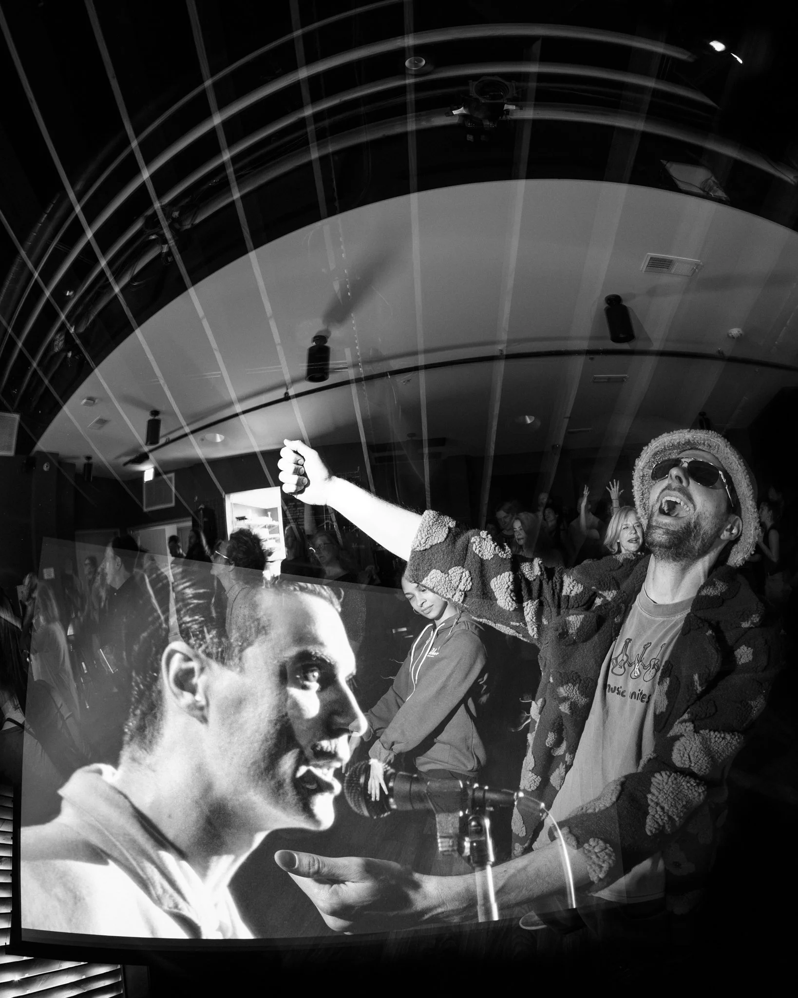 A black and white photo of a lively party scene with a man wearing sunglasses and a hat singing into a microphone, and a large screen displaying his face, in a crowded room.