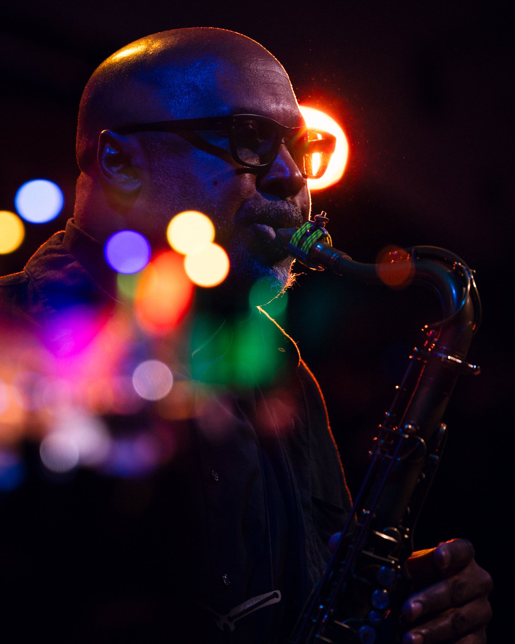 A man with glasses playing a saxophone surrounded by colorful bokeh lights in a dark setting.
