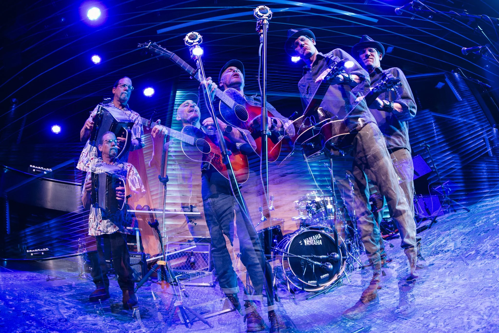 Multiple musicians performing on stage with guitars, drums, and accordion under blue stage lights in a concert venue.