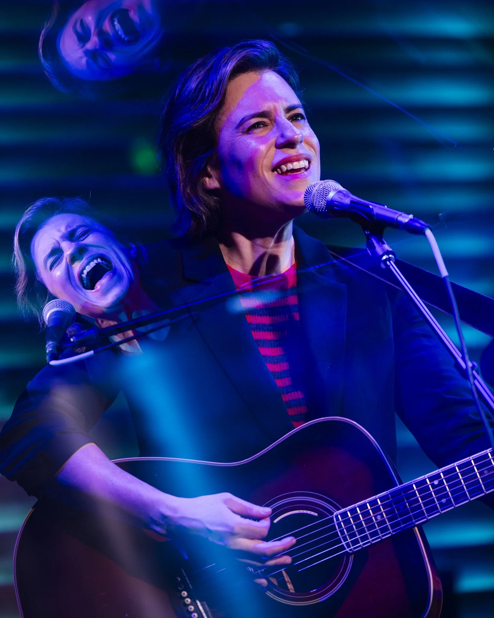 A woman with short dark hair playing an acoustic guitar and singing into a microphone, with a superimposed, larger image of her singing and smiling.