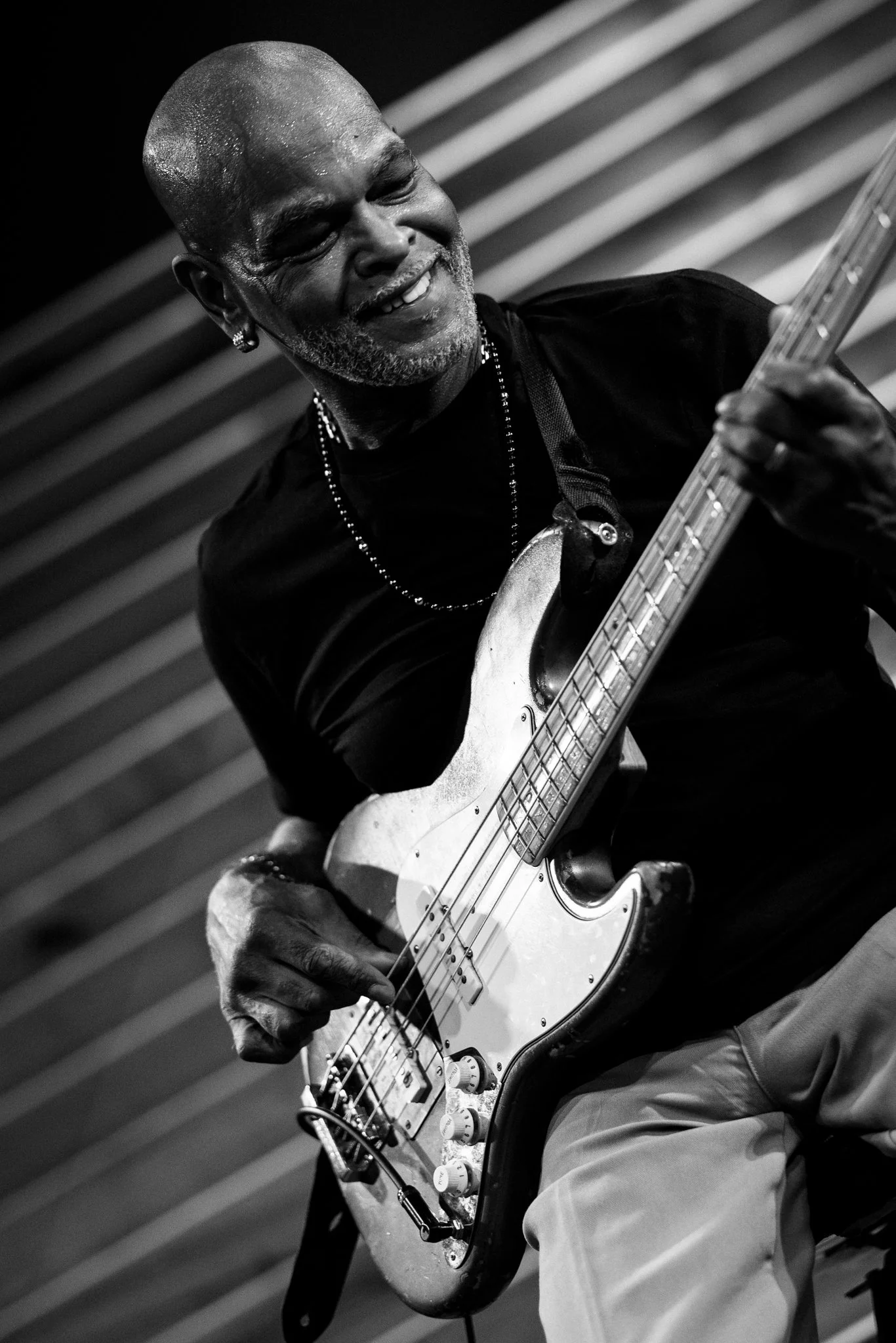 A smiling Black man with a bald head and gray beard playing an electric bass guitar.