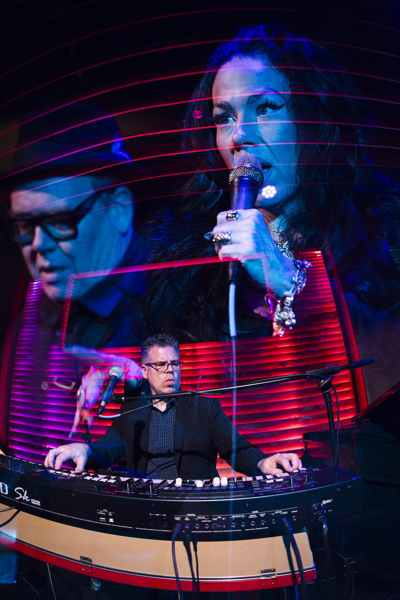 Two musicians performing on stage with a large video projection behind them. The woman is singing into a microphone, and the man is playing an electronic keyboard.