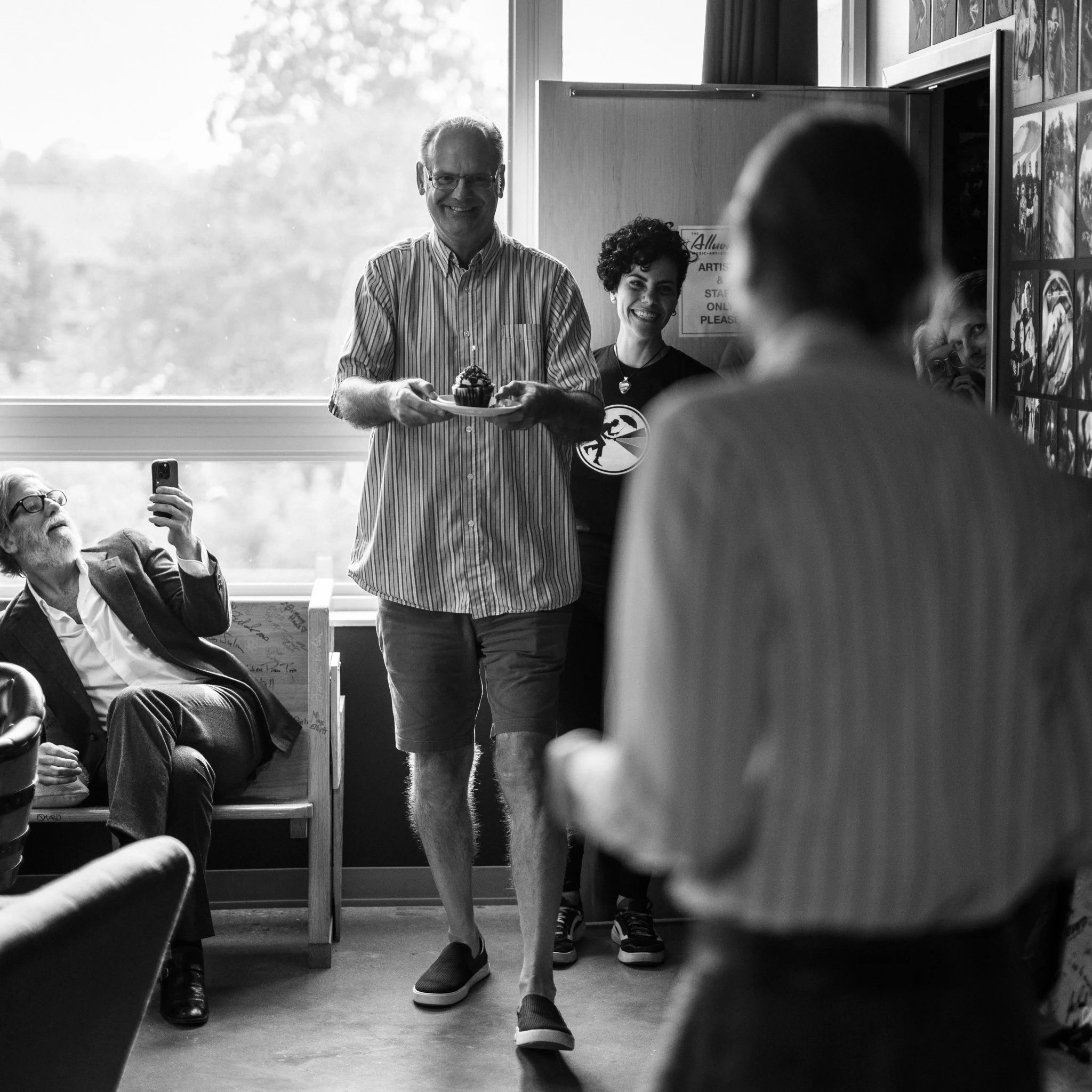 A man with glasses and a striped shirt holding a plate with a cupcake, smiling. A woman with curly hair and a black shirt stands behind him, smiling. Several seated and standing people are in the background, with some taking photos.
