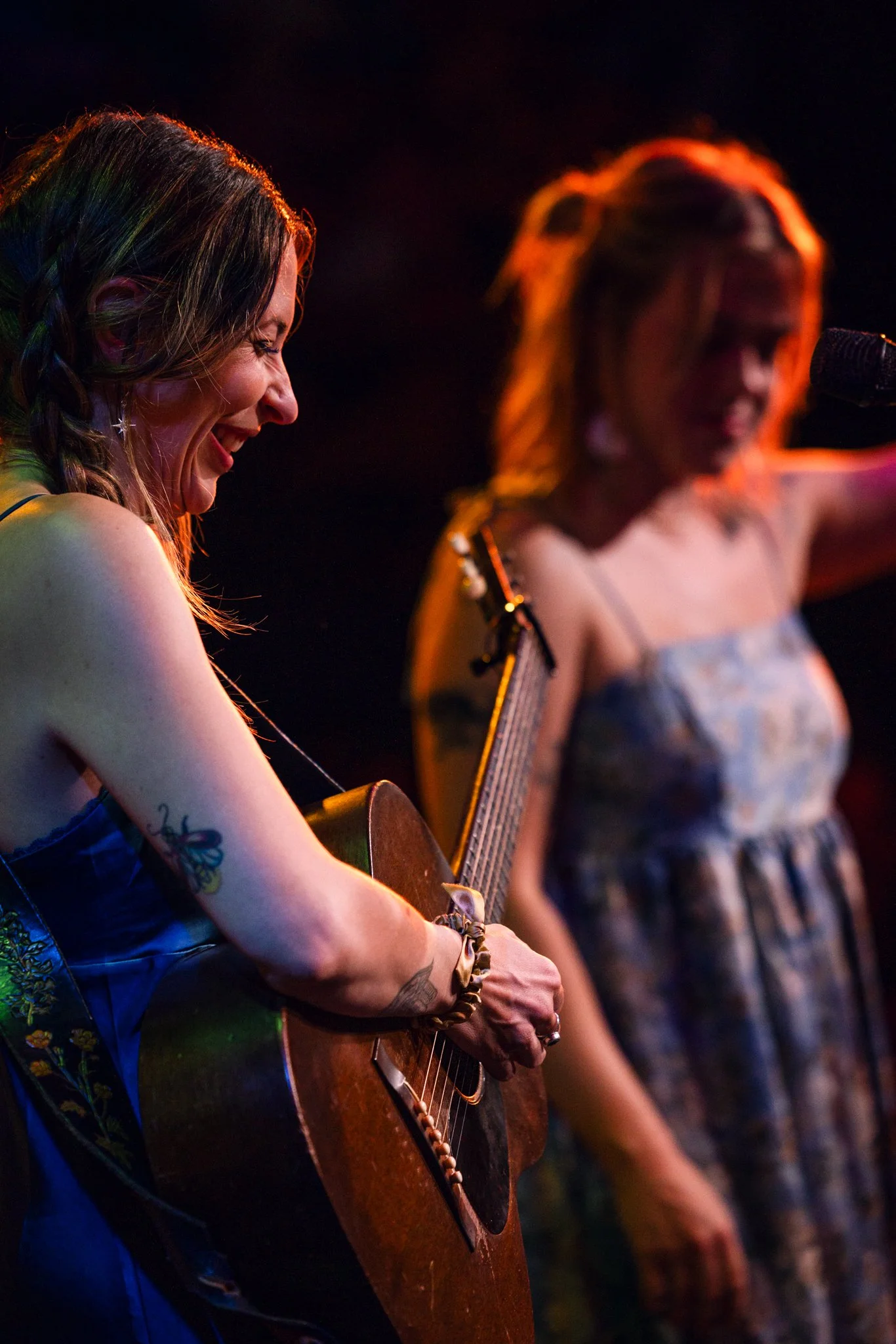 Two women performing on stage, one playing guitar and the other singing into a microphone, both smiling and enjoying the moment, under warm stage lighting.
