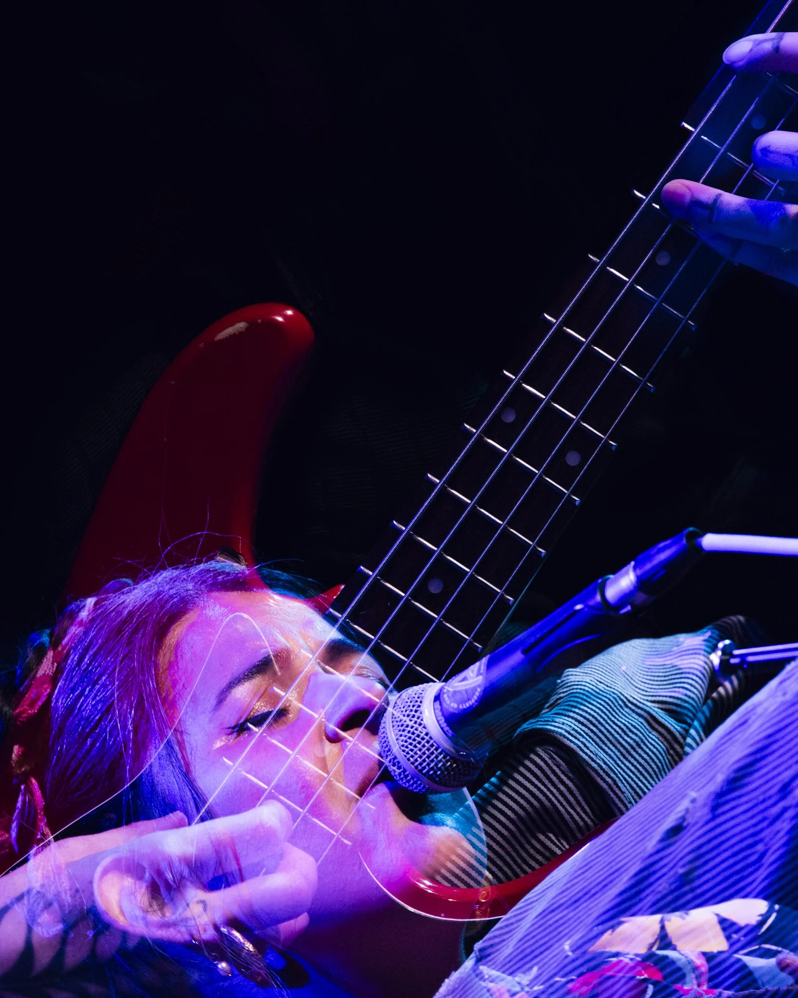 A woman singing into a microphone while playing an electric guitar on stage, with colorful stage lighting.