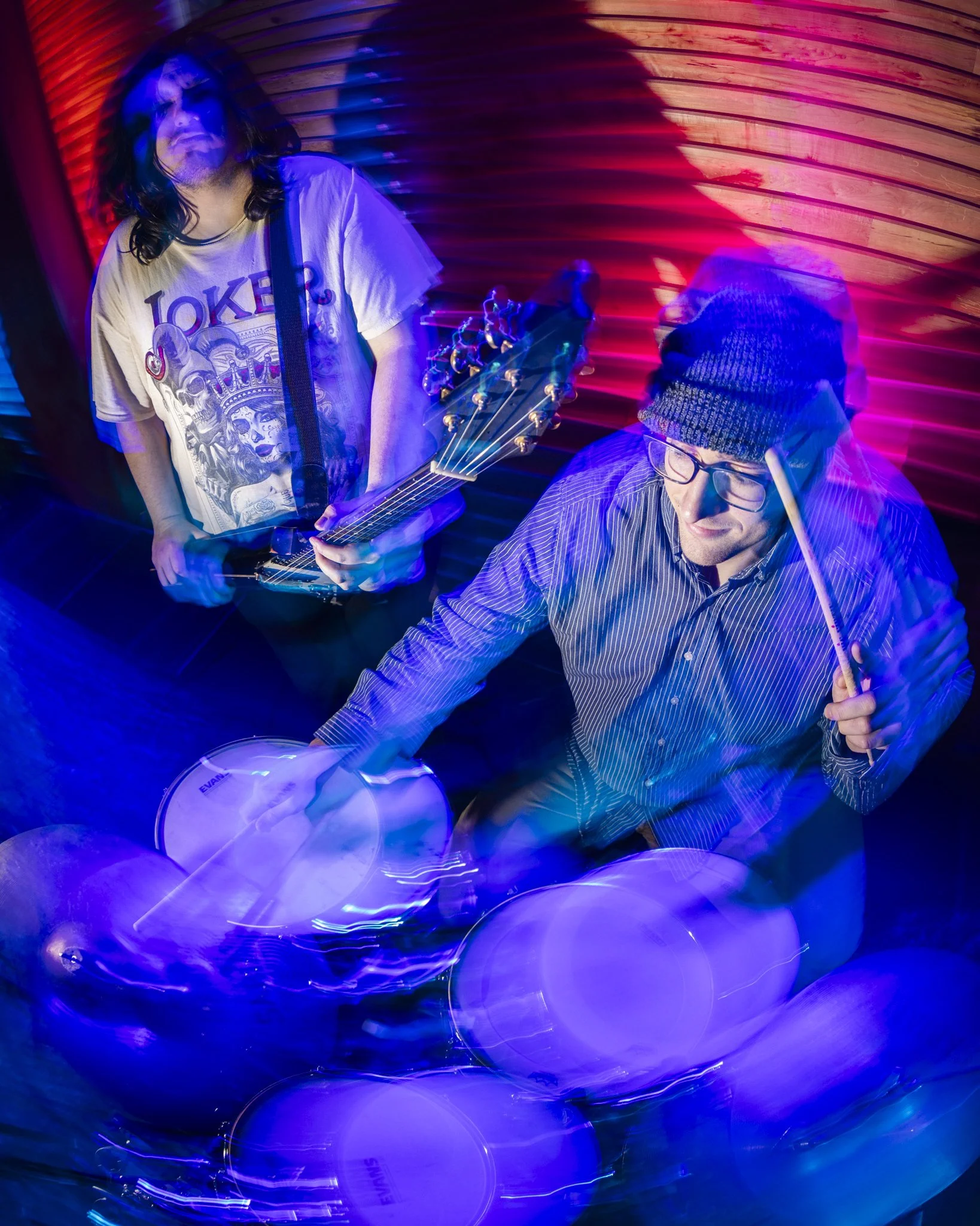 Two musicians, one playing an electric guitar and the other playing drums, in a space with vibrant blue and purple lighting and a wooden wall background.