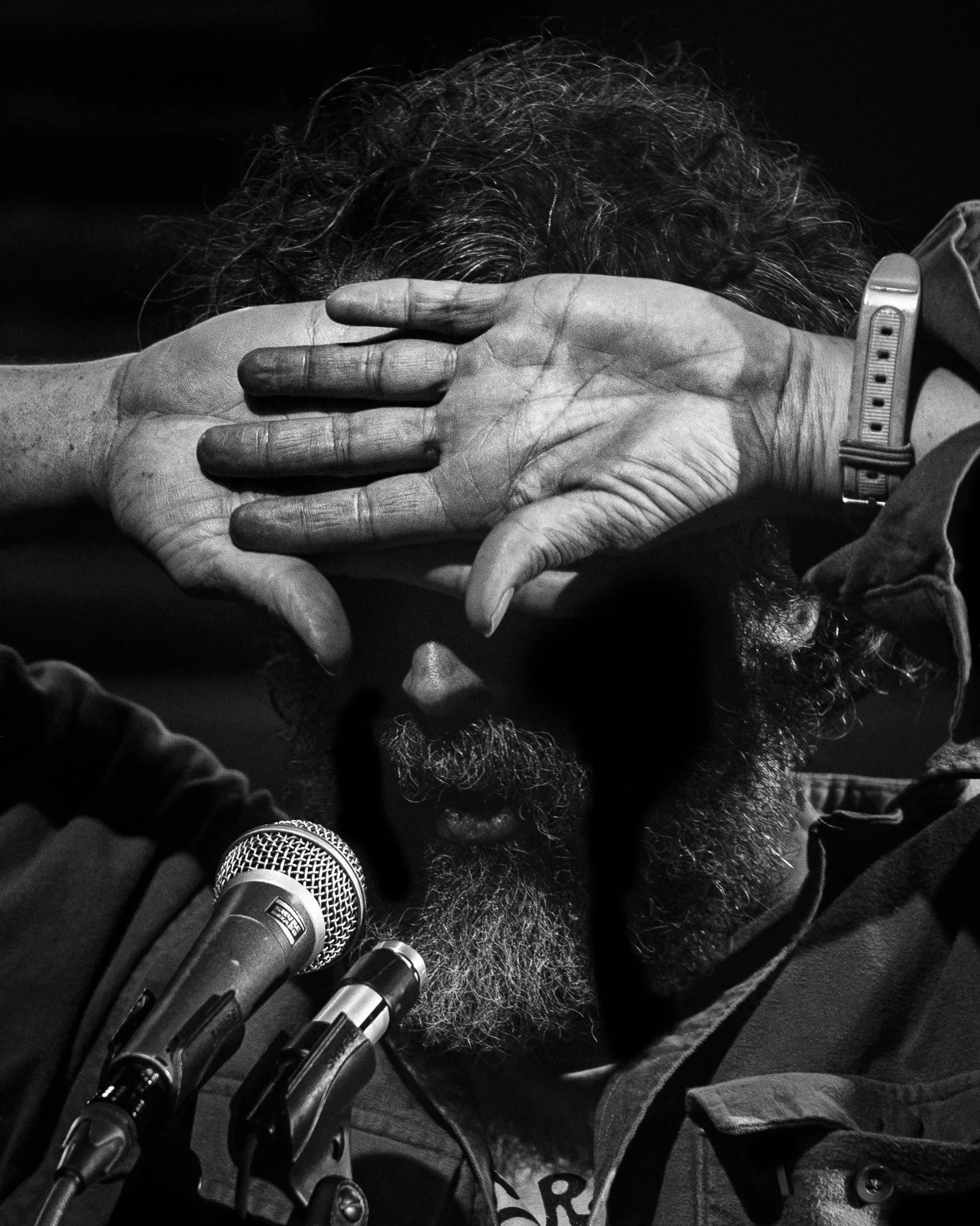 A man with curly hair and a beard covering his face with his hand, sitting in front of multiple microphones.