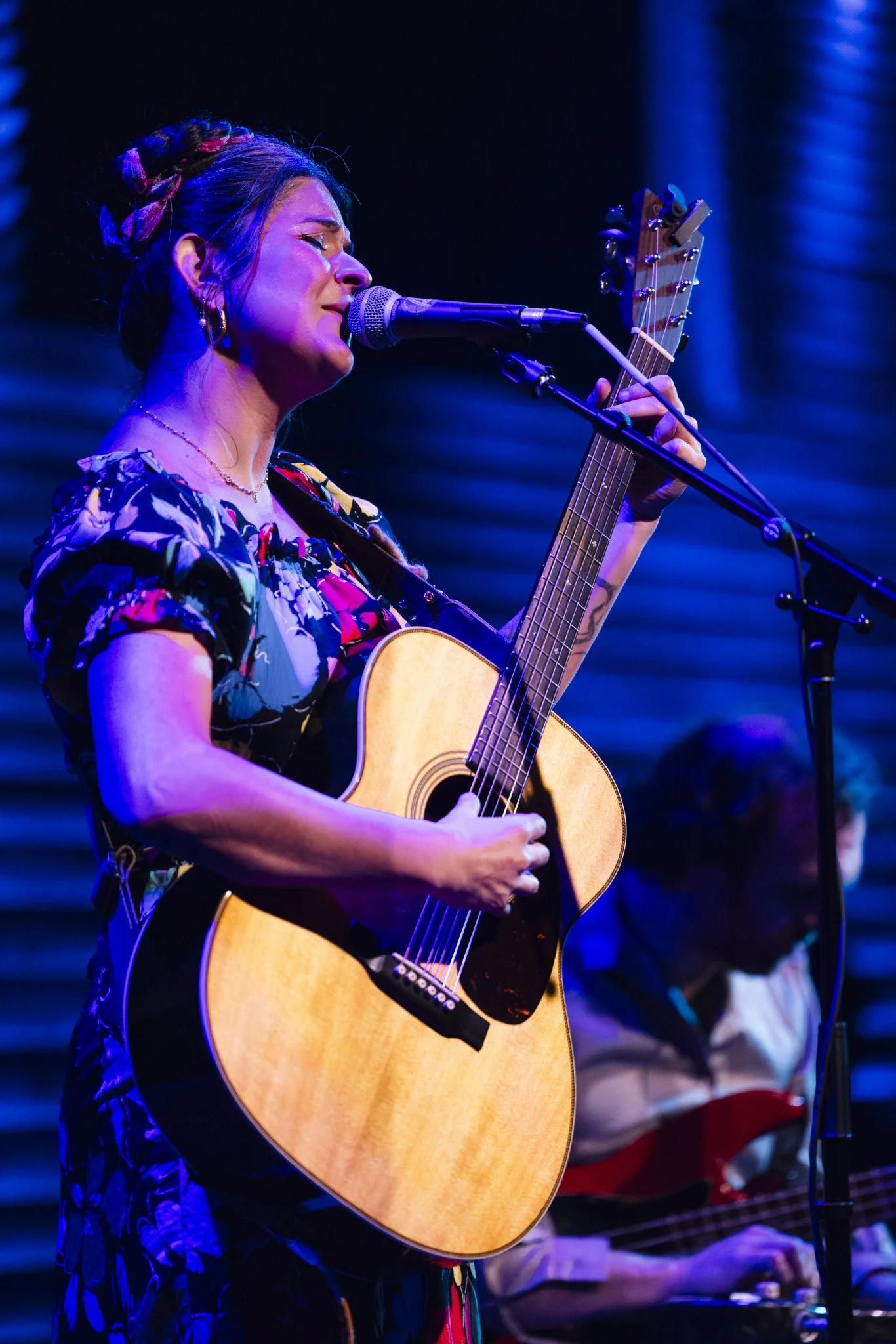 A woman singing and playing an acoustic guitar on stage, with a man in the background playing an electric guitar, under blue stage lights.