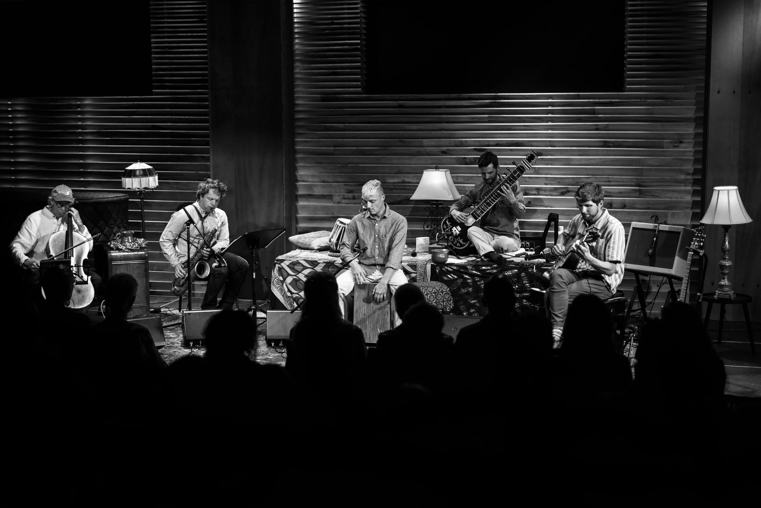 A black and white photo of a musical performance with five musicians on stage. They are playing various instruments including a guitar, saxophone, violin, and percussion, in a cozy, warmly lit setting with lamps and wooden walls. The audience is visi