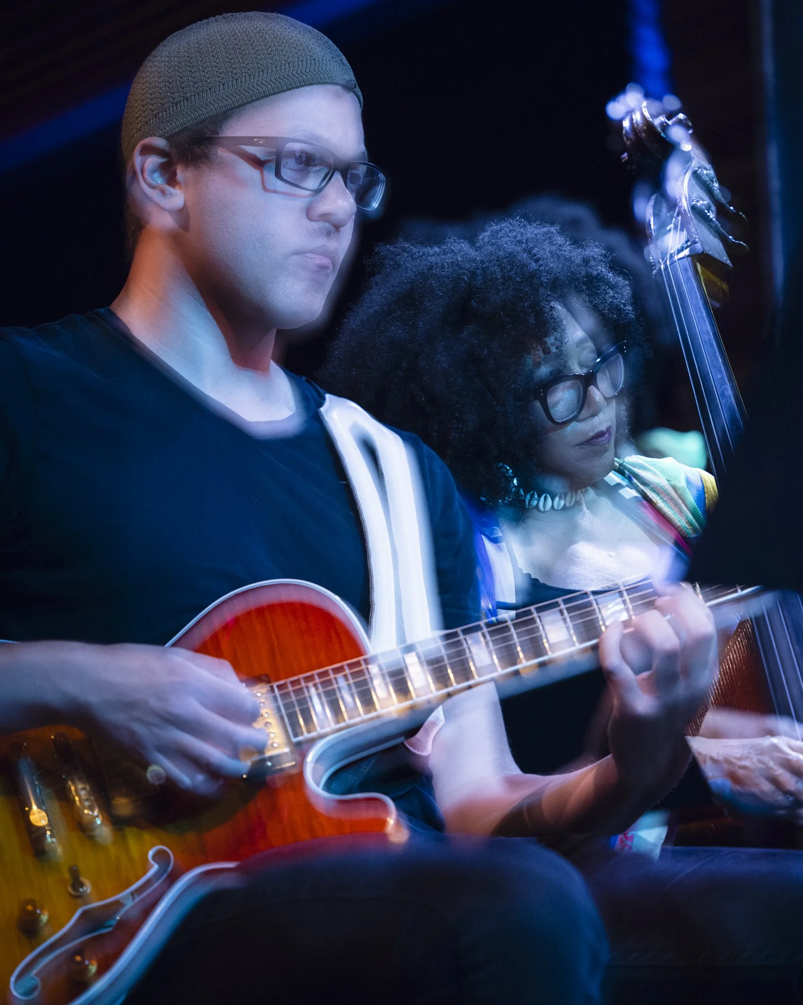 Two musicians, a man and a woman, playing guitars in a dark, blue-lit setting. The man has glasses, a beanie, and a black shirt. The woman has curly hair, glasses, and is wearing colorful clothing. The man is playing an electric guitar, while the wom