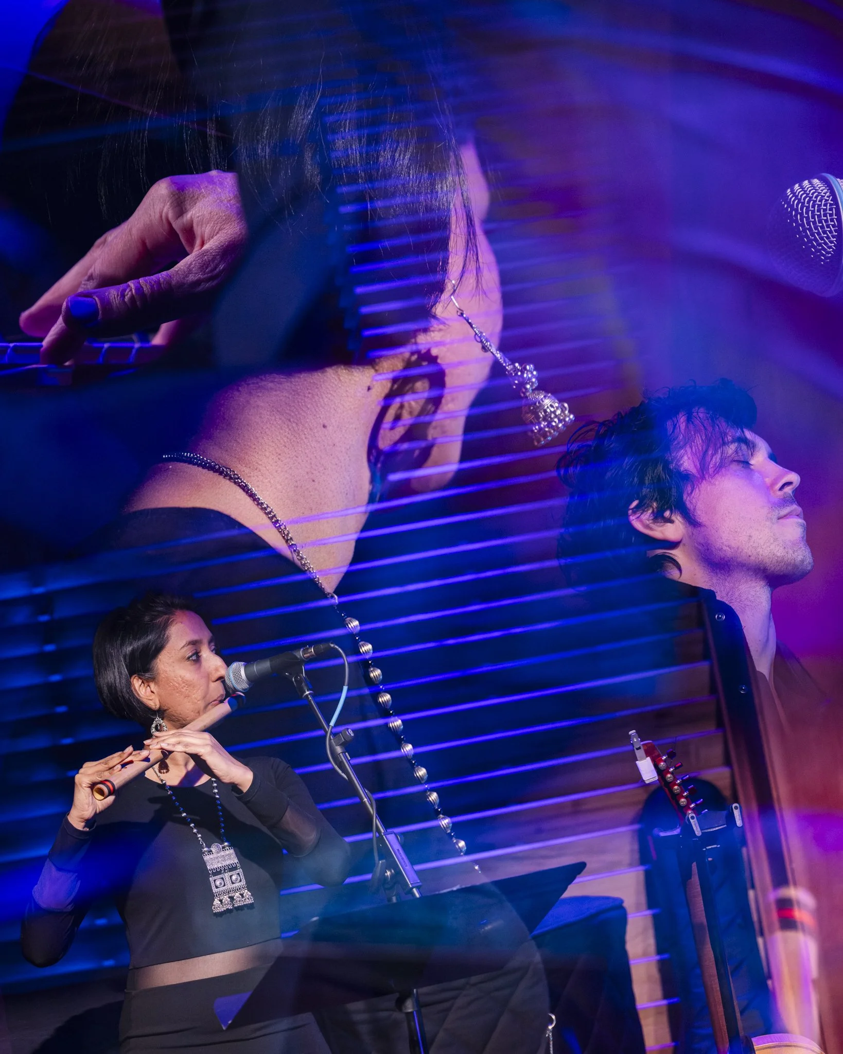 A woman playing a flute on stage with a man singing in the background, and blue and purple lights creating a layered, artistic effect.