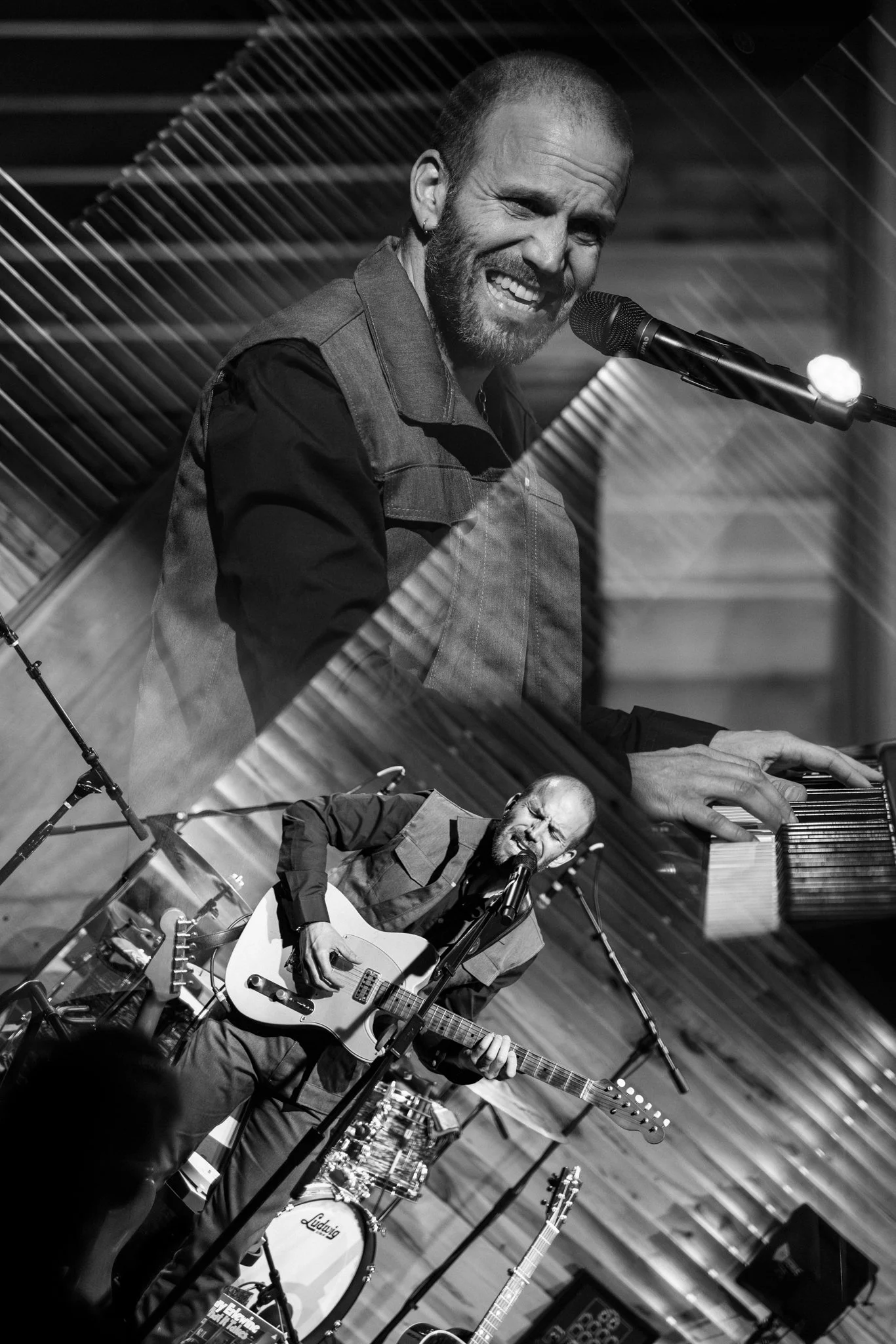 A black and white photo of a man singing and playing guitar, seen through fragmented glass with a pianist and drums in the background.