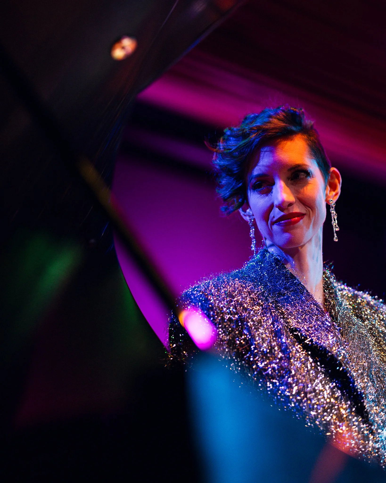 A woman dressed in a sparkling, glittery top with colorful stage lighting, gazing thoughtfully towards the right.