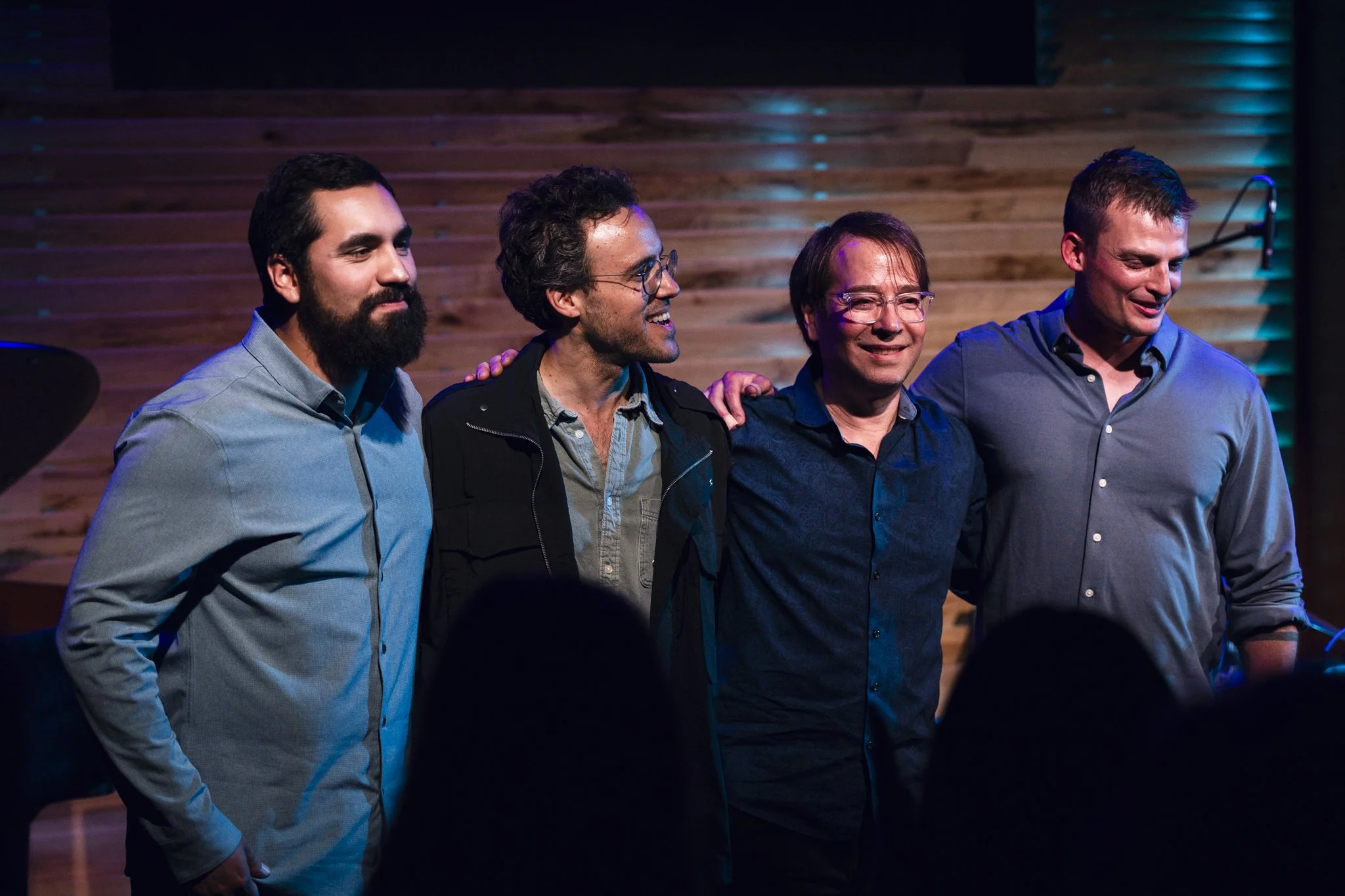 Four men standing together, smiling, with one man placing his hand on another's shoulder, in front of a wooden wall, in a dimly lit room.