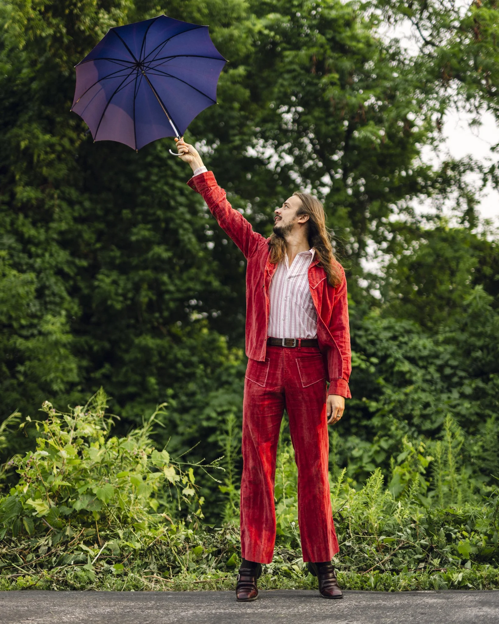 A man with long hair and a beard wearing a red velvet suit and moss green boots, standing outdoors on a paved road, holding a purple umbrella and looking up at it with a smile, surrounded by lush green trees and bushes.
