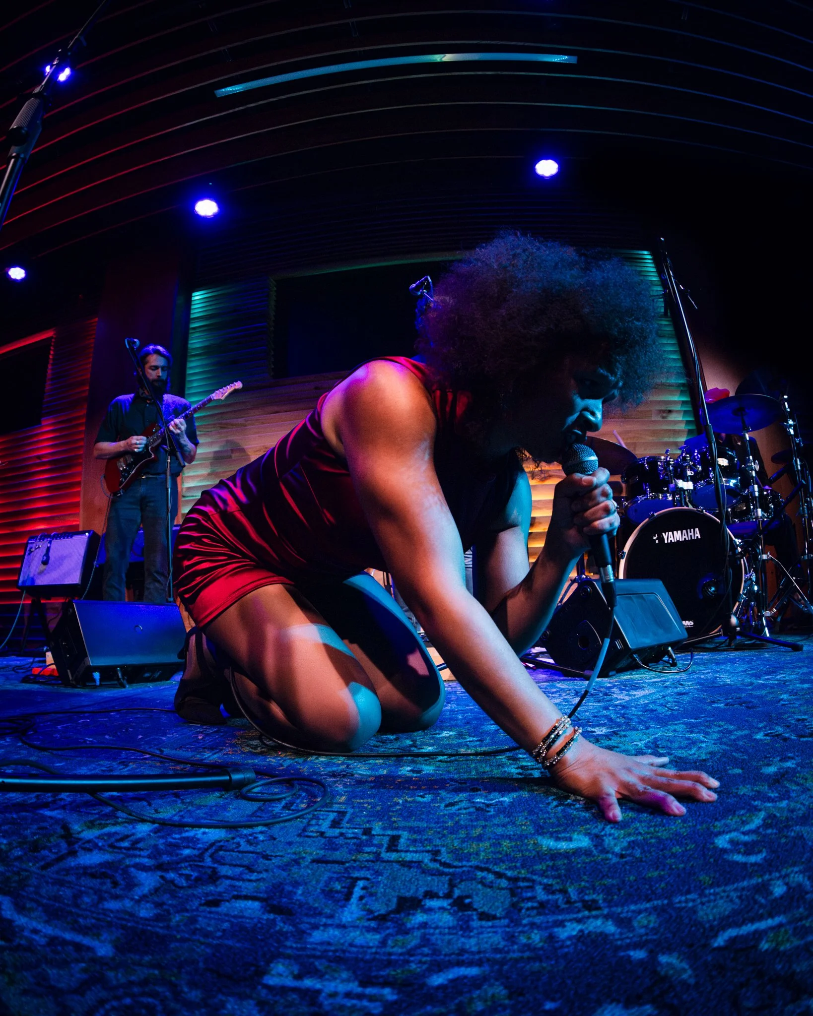 Musician kneeling on stage, singing into a microphone with a band in the background.